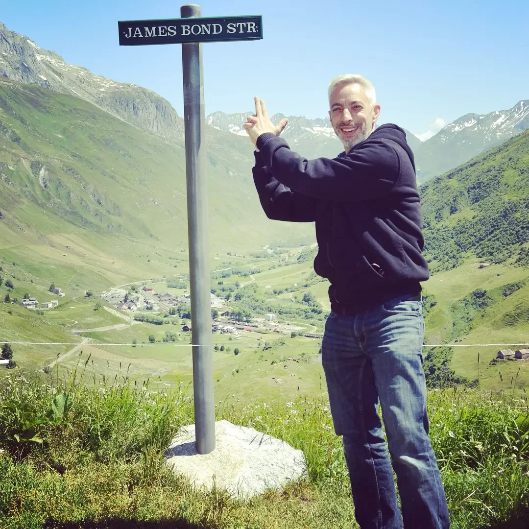 A man with short blond hair and a beard standing outdoors in a mountainous area, smiling and pointing at a street sign that reads 'James Bond Str.' The landscape includes green hills, scattered houses, and snowy mountain peaks under a clear blue sky.