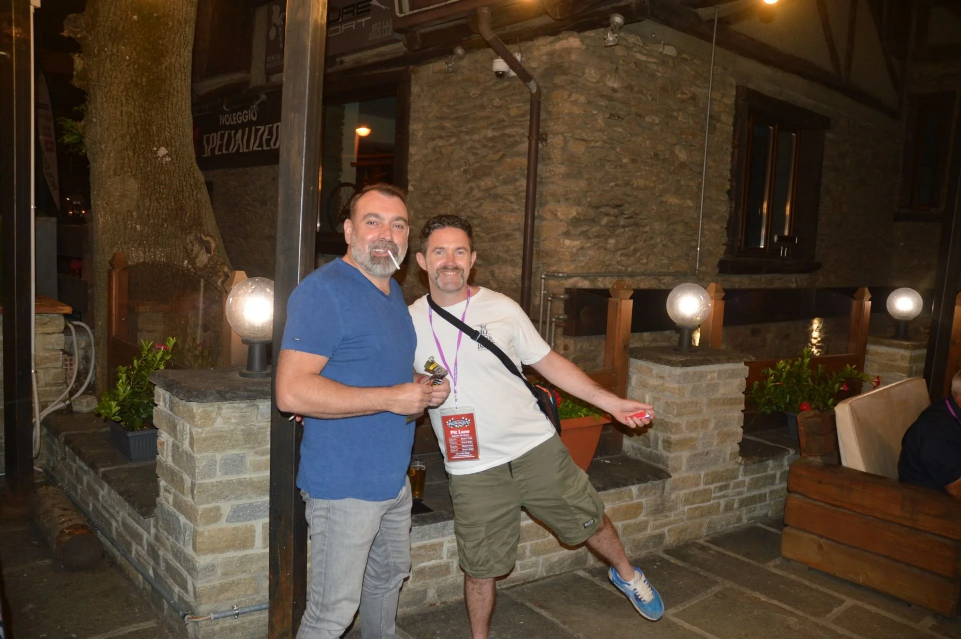 Two smiling men standing inside a rustic restaurant with stone walls, plants, and globe-shaped lights. One man is wearing a blue t-shirt and gray pants, holding a cigarette. The other man is wearing a white t-shirt and khaki shorts, with a lanyard ar