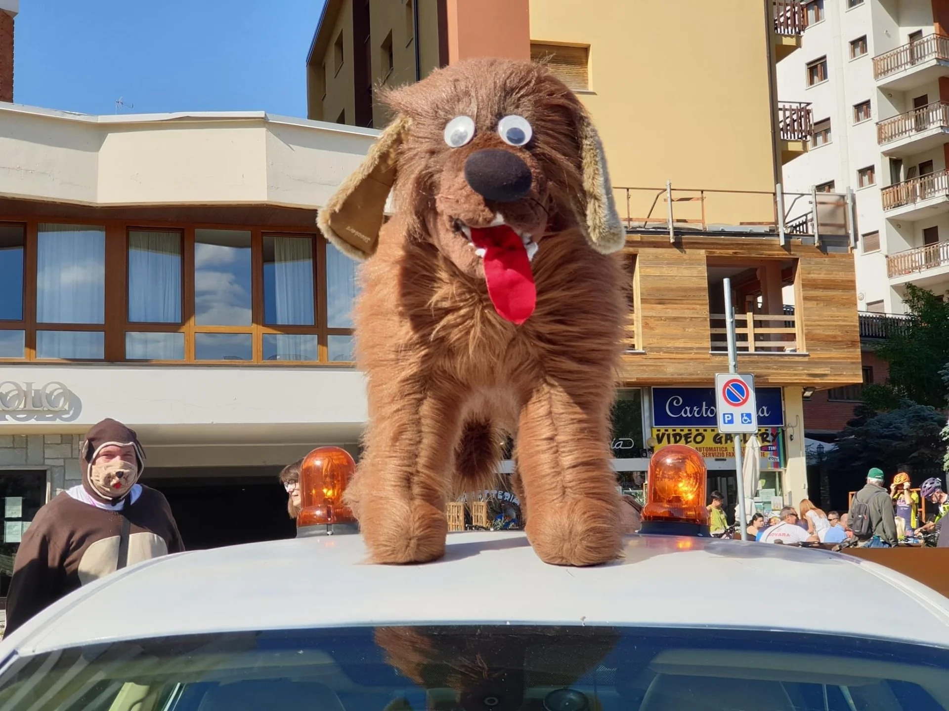 A person dressed in a dog costume celebrates on top of a vehicle at a parade or festival, with buildings and onlookers in the background.