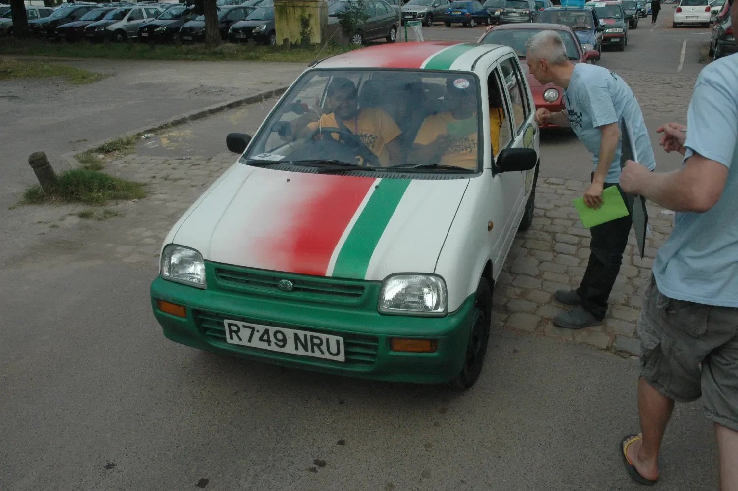 A small white car with a green front bumper and a red, white, and green stripe on the hood, resembling the Italian flag, parked on a cobblestone surface near a parking lot. Two men are interacting with the driver inside the car during daytime.