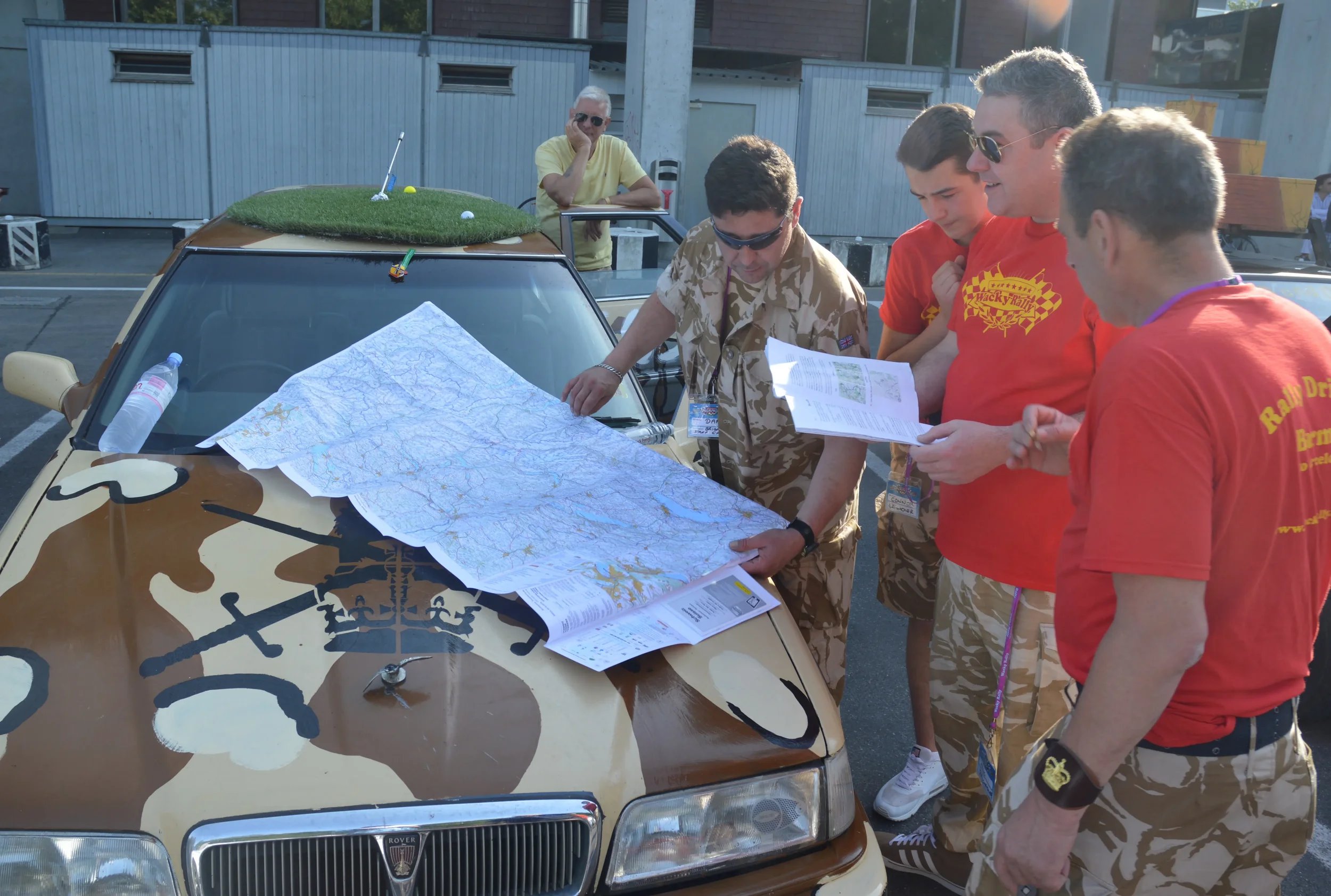 Group of five men studying a large map spread out on the hood of a camouflage-painted car with a 1980s design, parked in a lot. One man in military-style uniform points at the map; the others, four in bright red T-shirts and one in sunglasses and a y