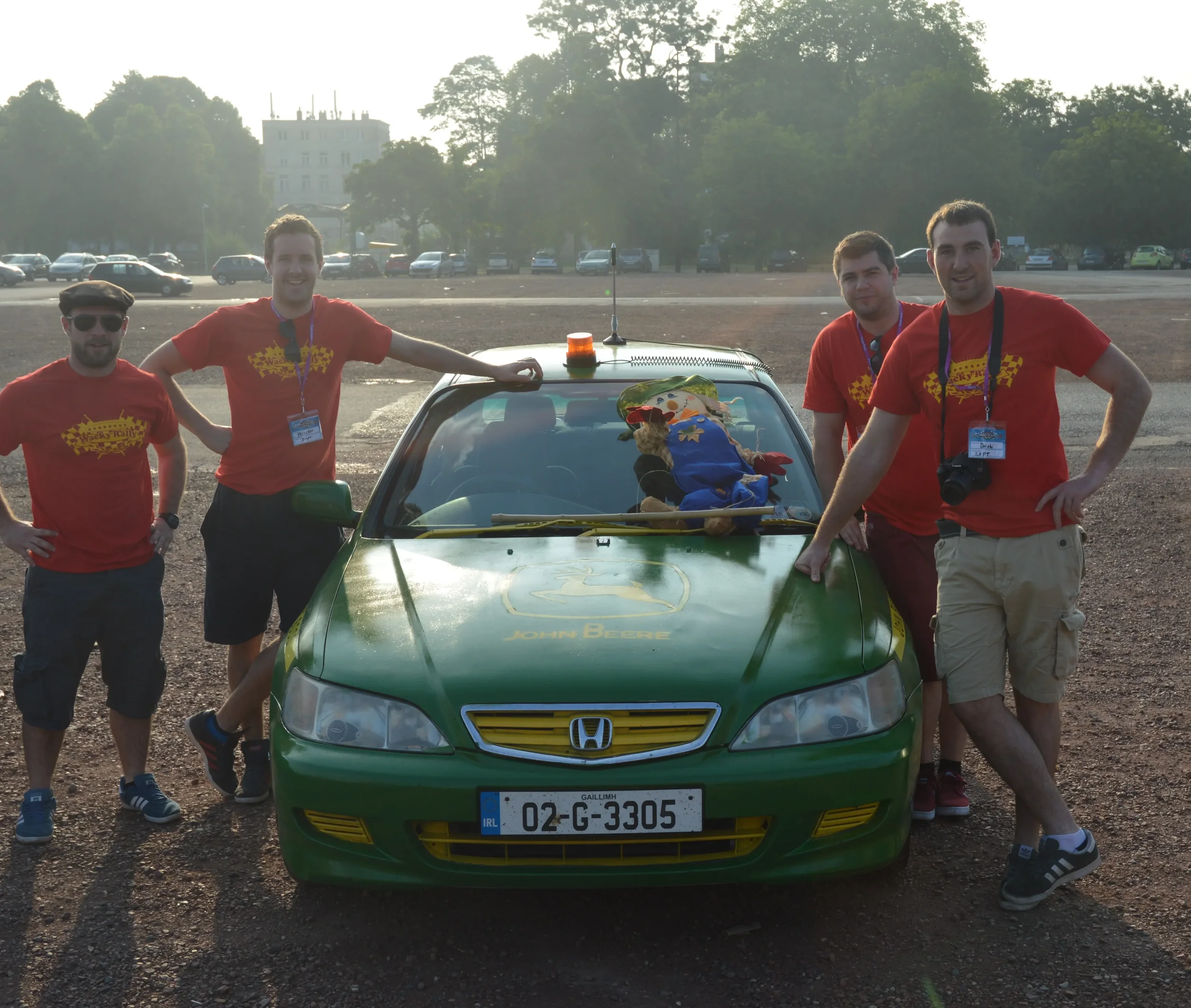 Four men standing around a green Honda race car, smiling, dressed in red t-shirts with yellow checkered flag designs, in an outdoor parking lot during daylight.