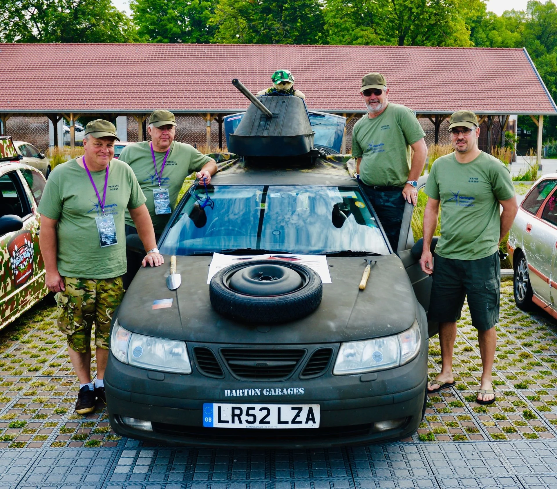 Group of four men standing around a car equipped with military and tank features, including a tank turret on the roof and a spare tire on the hood, in an outdoor parking lot.