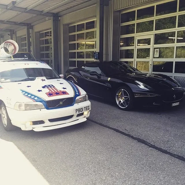 A police car and a black Ferrari are parked side by side outside a building with large garage doors.