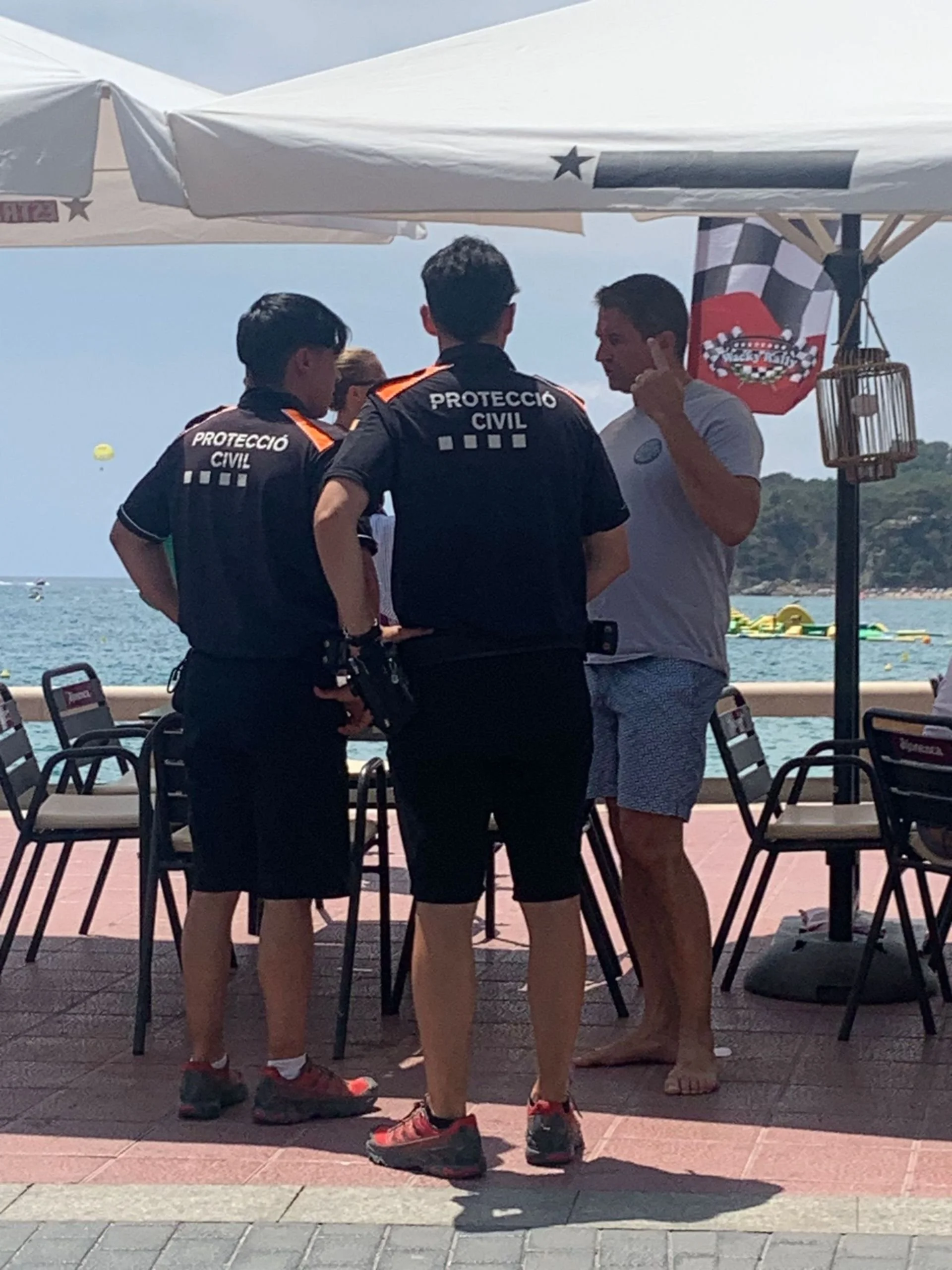 Two civil protection officials and a man in casual clothes having a conversation on a seaside patio, with chairs, umbrellas, and the water in the background.
