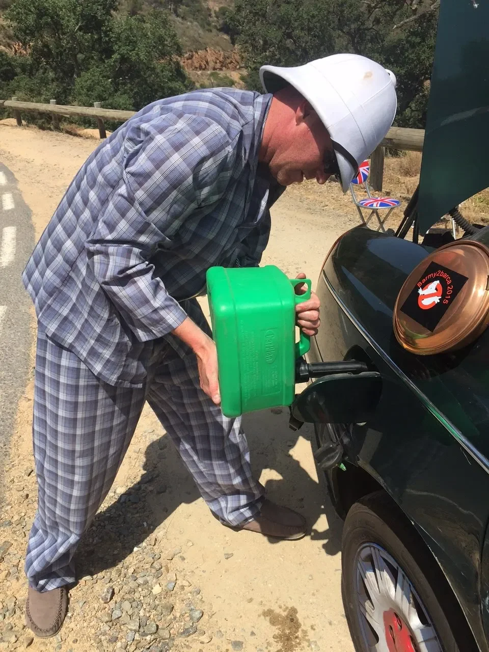A man dressed in plaid pajamas and a large white hat refueling a black car with a green gas container outdoors on a dirt road.