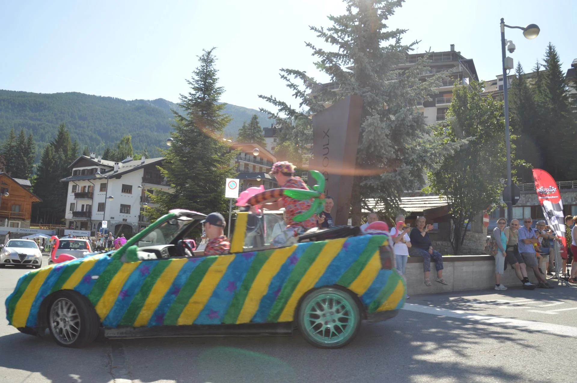 A parade car decorated with colorful stripes and purple stars driving on a street, with people watching from the sidewalk and bunting flags in the background, surrounded by trees and buildings, against a mountainous landscape.