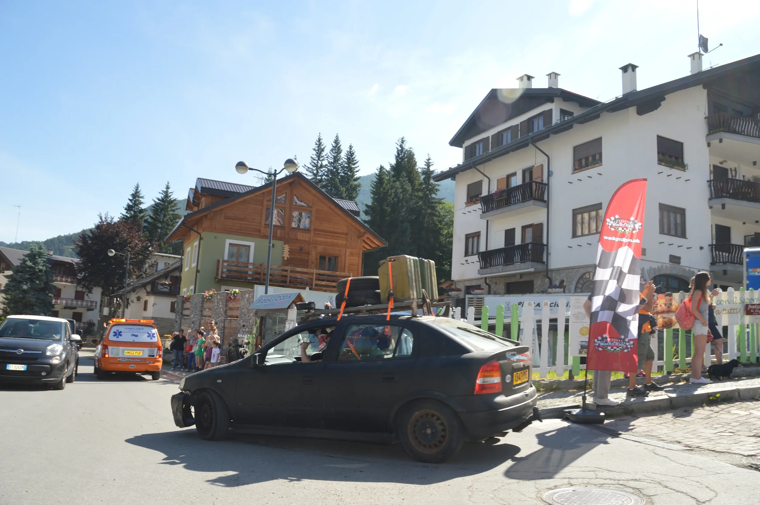 A black car with luggage on top parked on a street in a village with people, an ambulance, and buildings in the background.