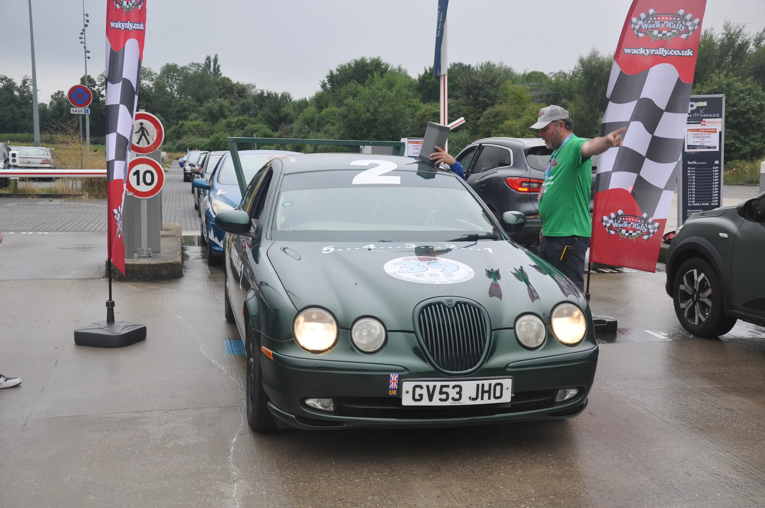 A dark green vintage car at the starting line of a rally event, with a man in a green shirt and cap pointing to the right and a person on the car's roof holding a clipboard. The car has a logo on the hood and a UK license plate, surrounded by red ral