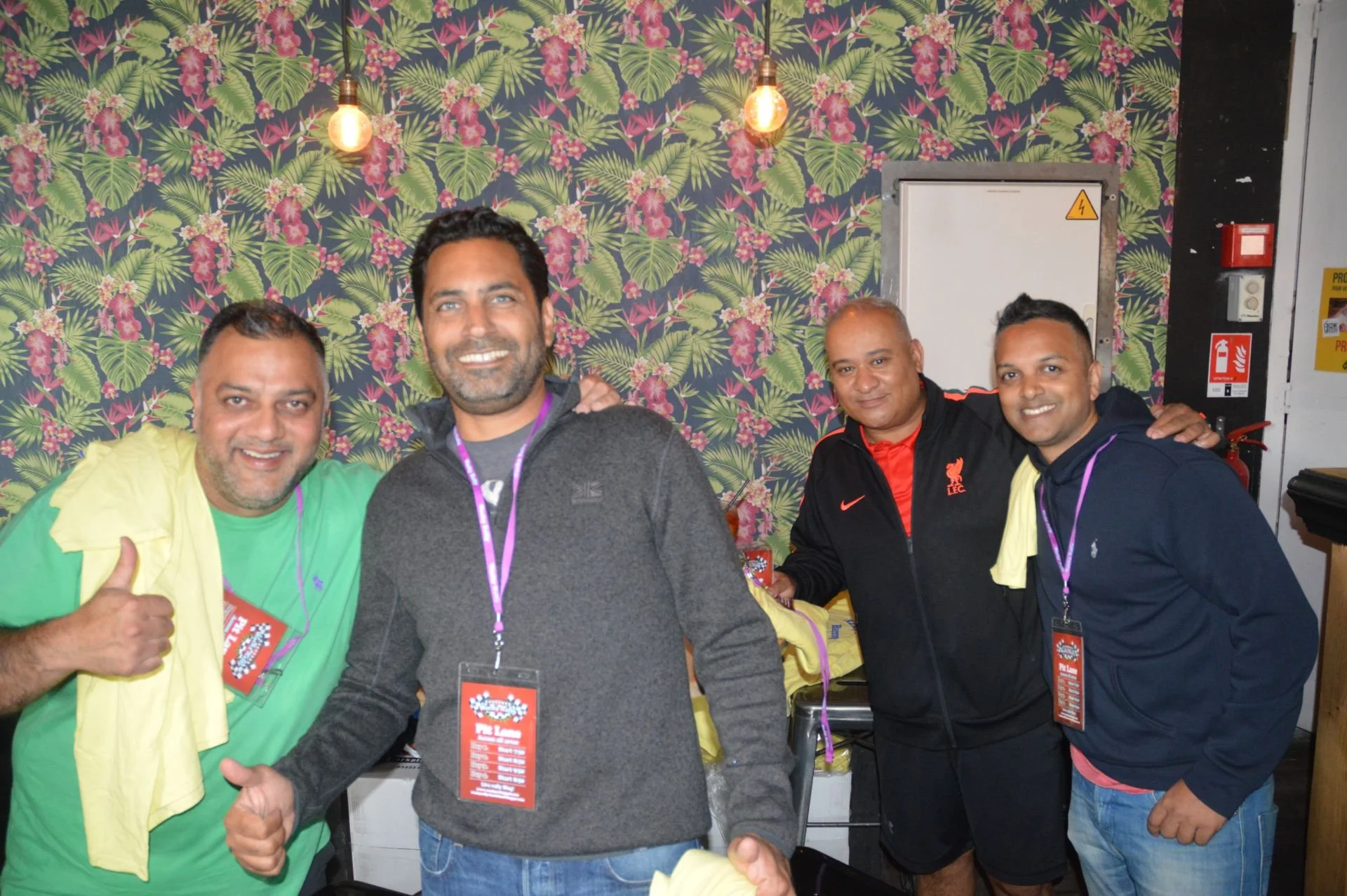 Four men standing together, smiling, with a floral wallpaper background. They are wearing casual clothes and event lanyards.