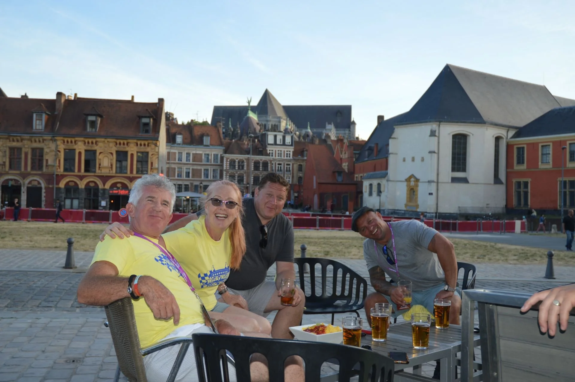 Four people sitting outdoors at a table with drinks, smiling and posing for the photo, with historic European-style buildings in the background.