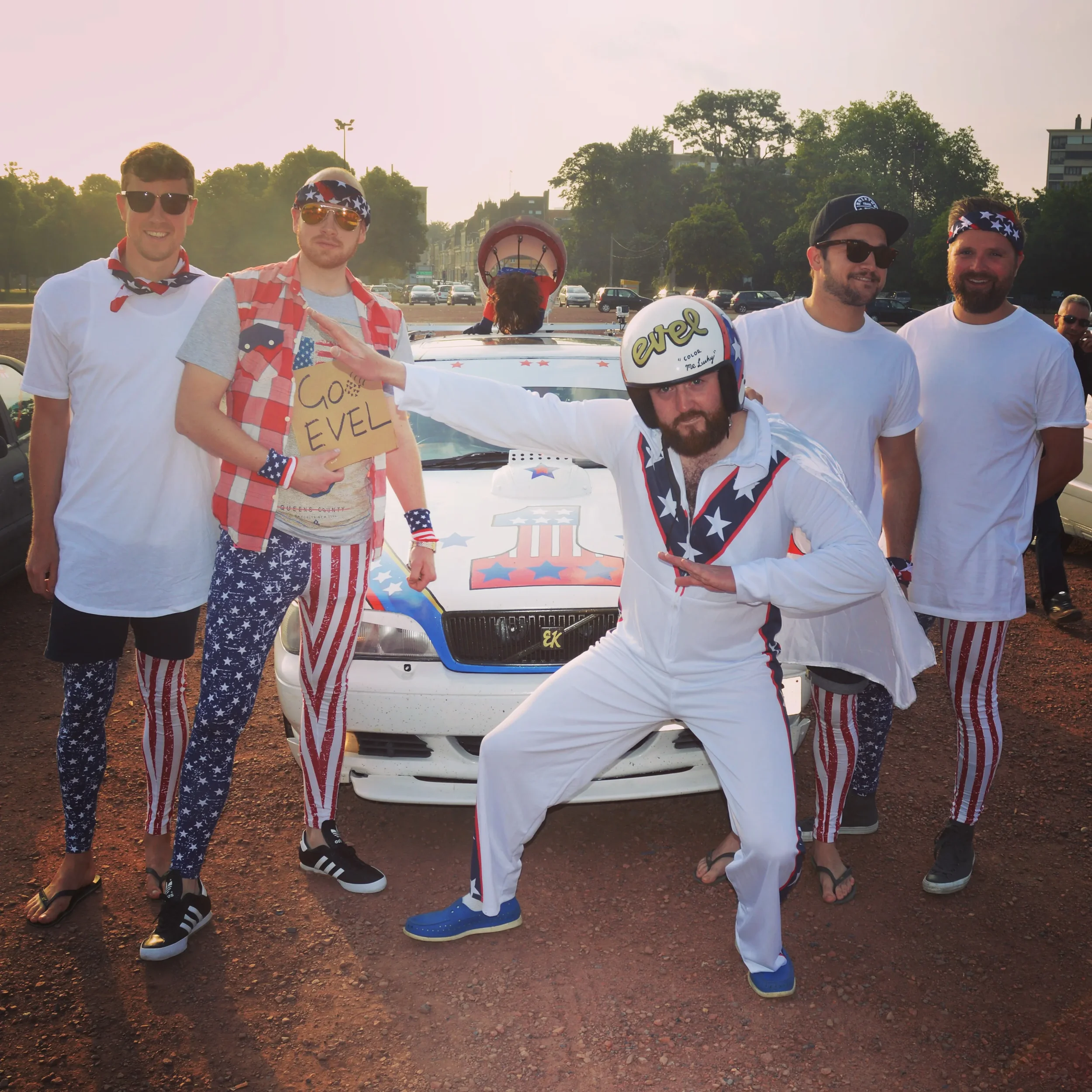 A group of six people dressed in patriotic American-themed clothing, posing in front of a decorated car with a large red, white, and blue emblem. They are wearing flag-themed pants, shirts, and accessories, and are celebrating an event related to the