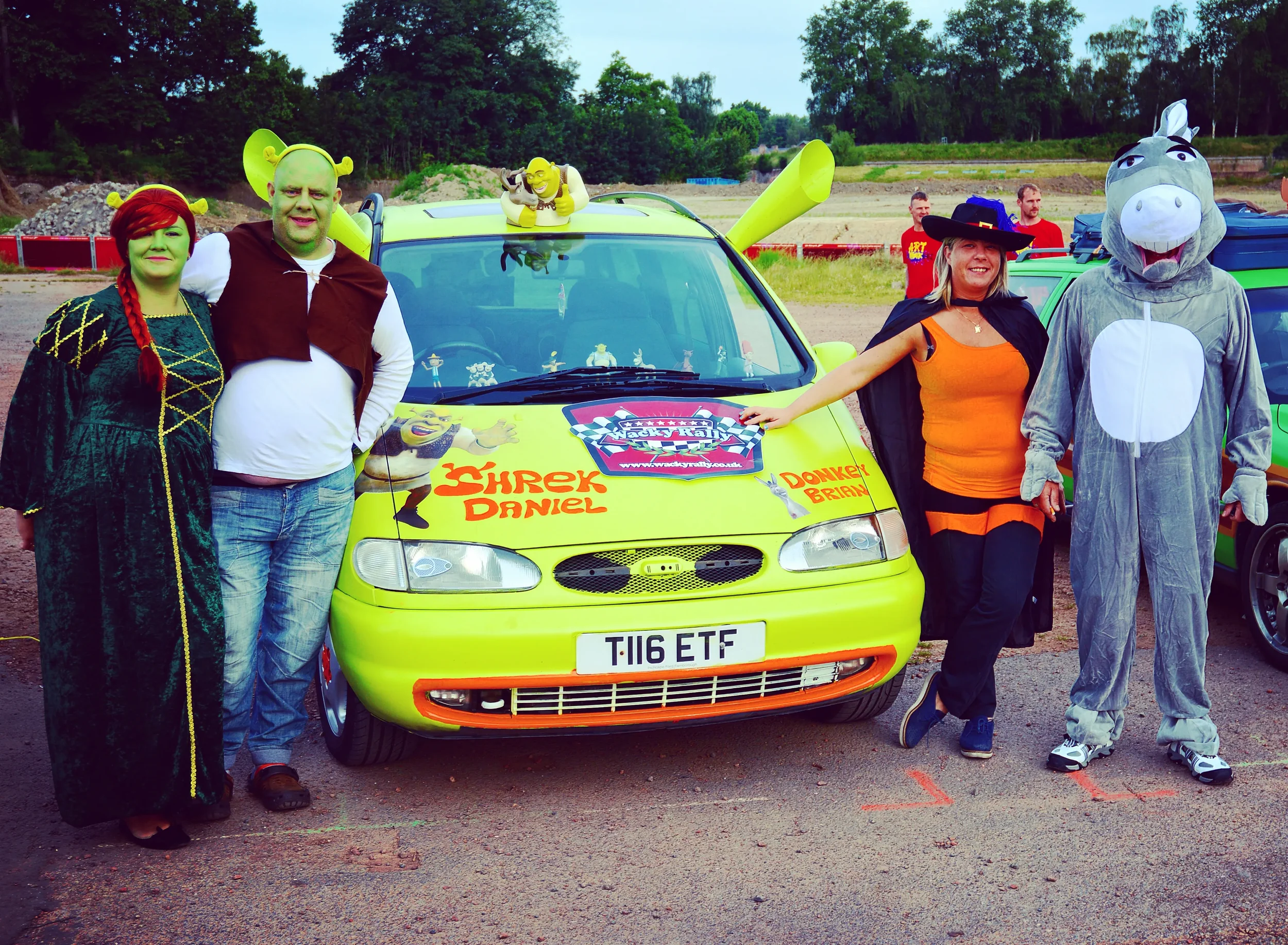 Group of people dressed in costumes standing in front of a yellow car with cartoon character stickers, including Shrek and Donkey, on the hood. The scene appears to be at a themed event or gathering.