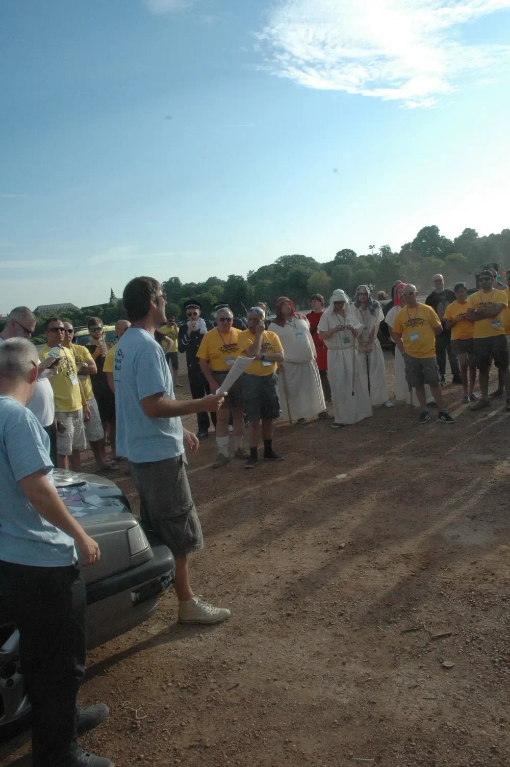 Group of people gathered outdoors, listening to a man holding papers, with some wearing yellow shirts and traditional Middle Eastern clothing, on a sunny day with trees and a building in the background.