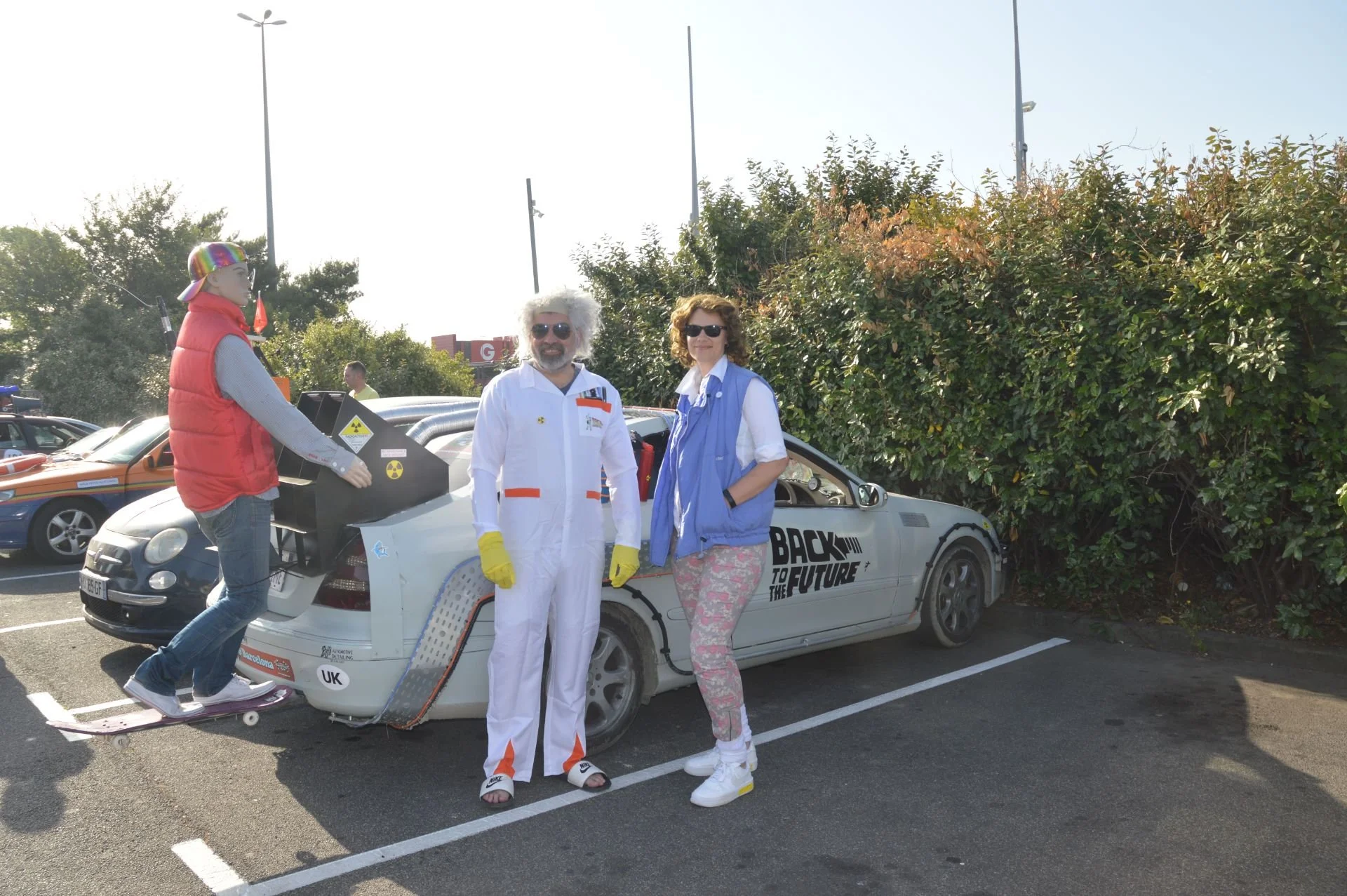 Three people posing in front of a DeLorean car with "Back to the Future" logo, one person on a skateboard near the car, two people standing beside it, all outdoors in a parking lot with cars and bushes in the background.