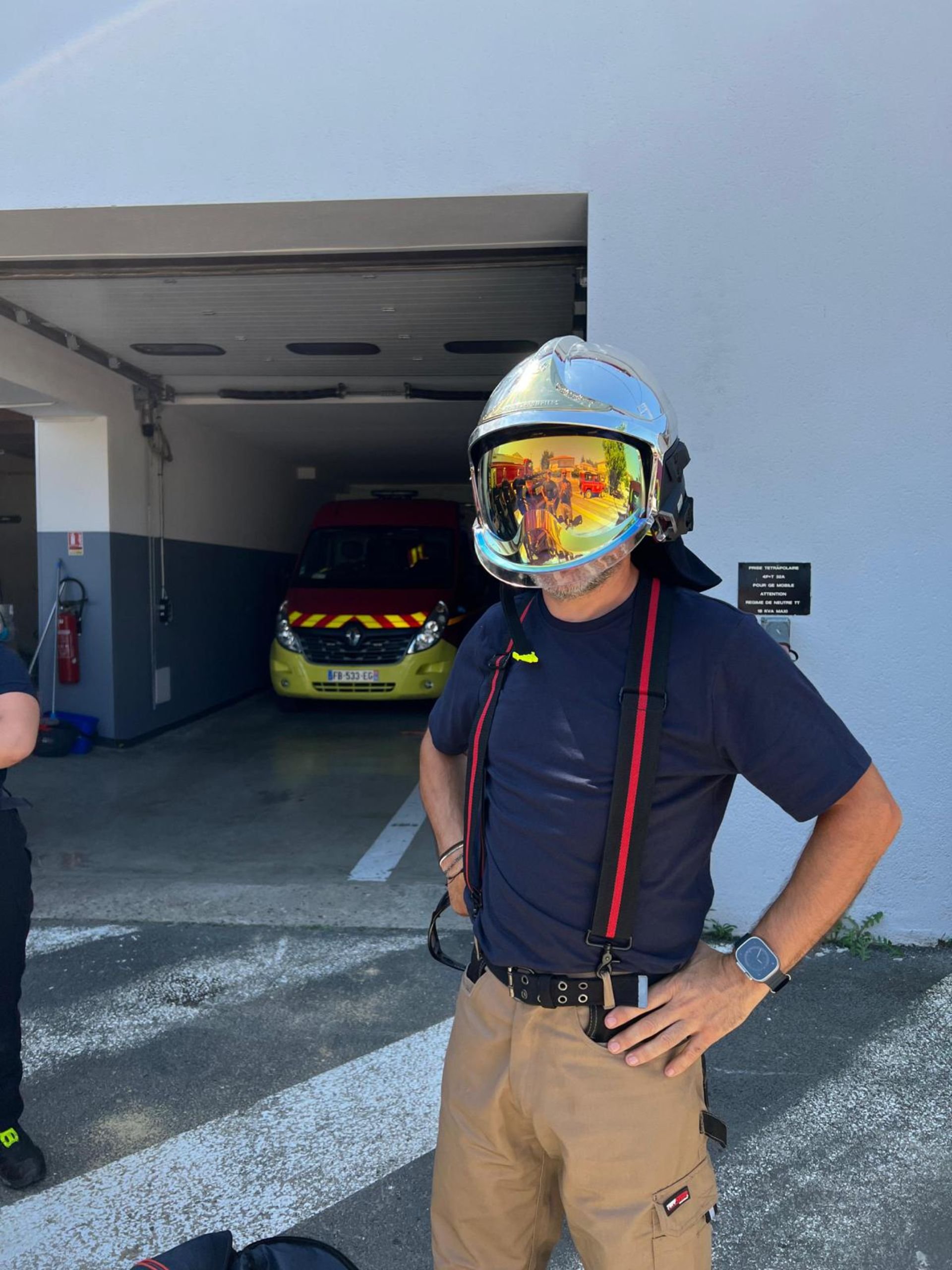 A firefighter standing outside near a parking garage, wearing a helmet with reflective visor, black T-shirt, khaki pants, and a wristwatch.