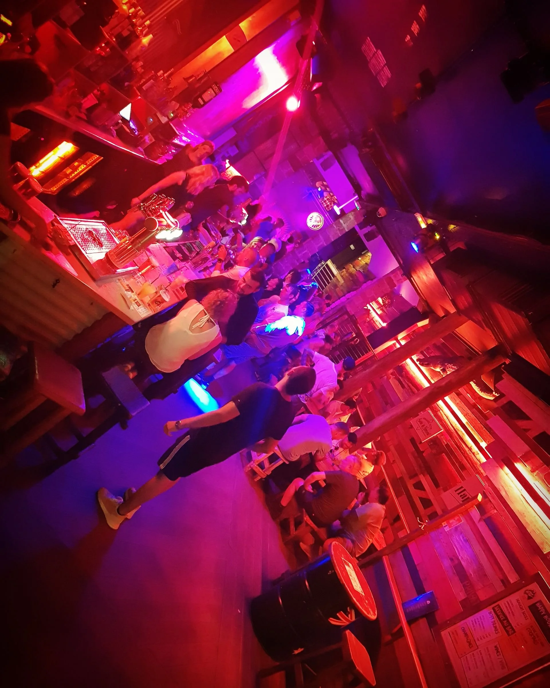 A crowded bar with people socializing and drinking, illuminated by colorful red, purple, and pink lighting, with bartenders serving drinks behind the counter.
