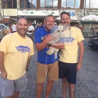 Three men standing outdoors holding a white dog with a clear plastic bag around it, in front of a Carrefour store.