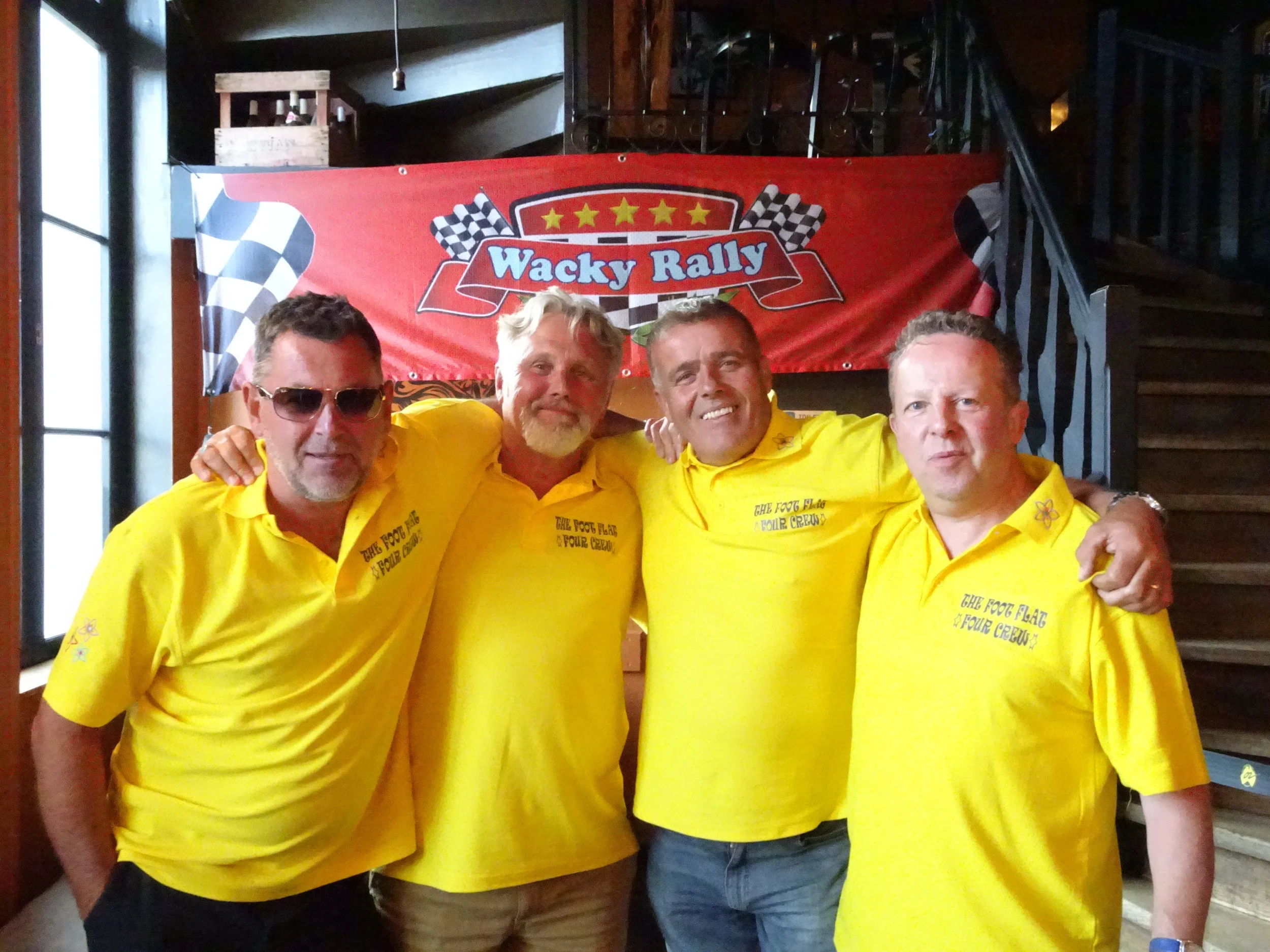 Four men dressed in yellow polo shirts posing together indoors, with a red banner reading 'Wacky Rally' and checkered flags in the background.