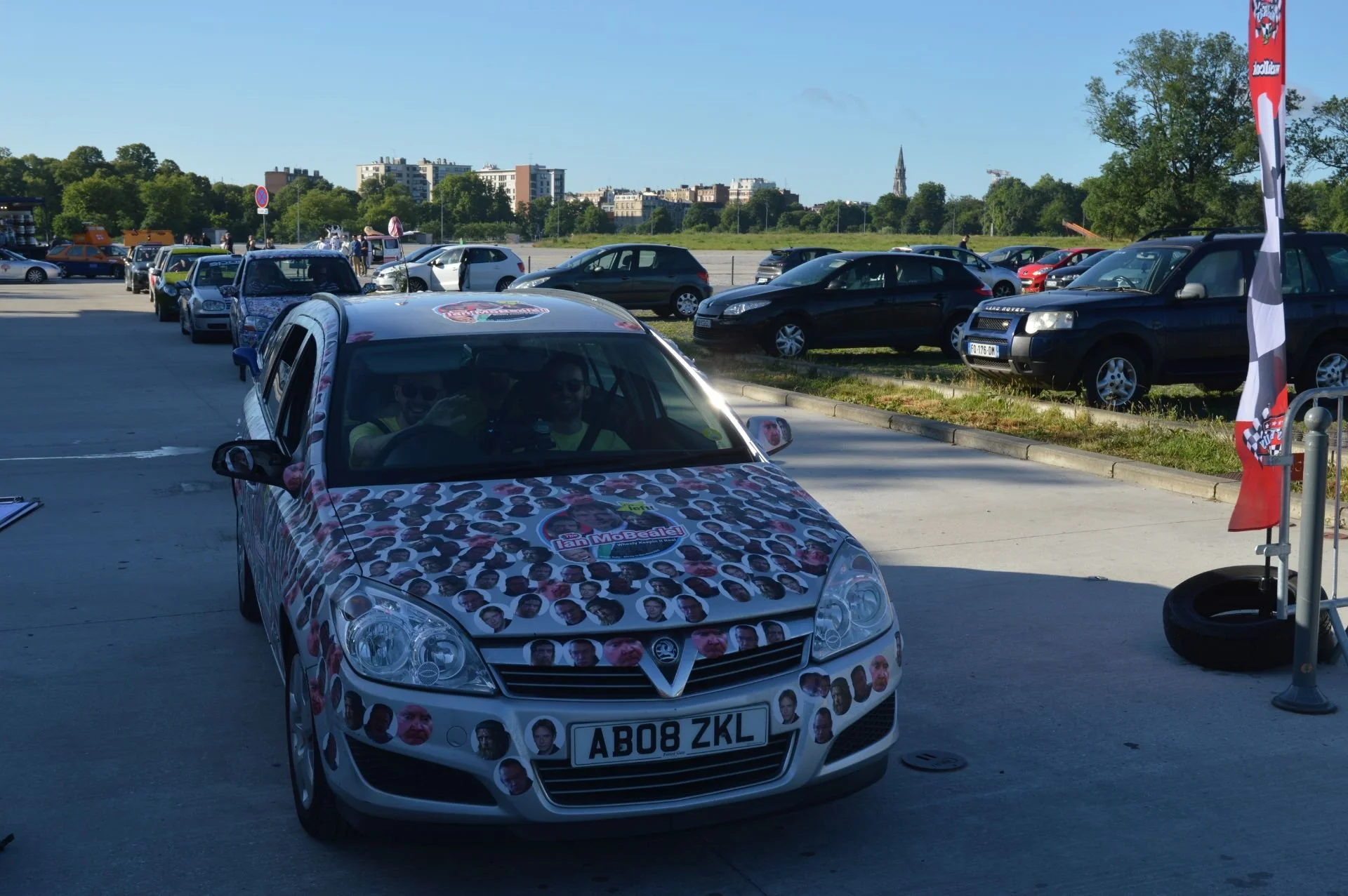 A white car decorated with numerous photos of people's faces and a logo reading "La MoBeaute" parked in a parking lot with other cars and trees in the background.