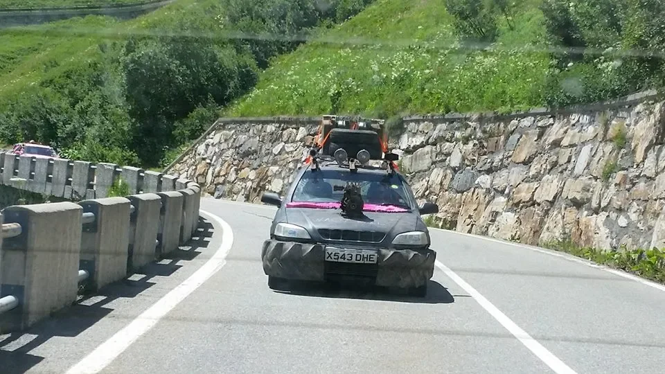 A gray car decorated with black tire treads and purple ribbon, with camera equipment and luggage on the roof, driving on a winding mountain road with protective barriers and rocky slopes.