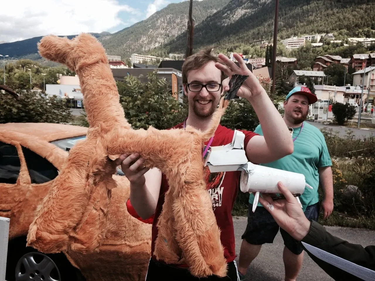Two people, one holding a large, plush, orange animal costume, the other smiling near them, outdoors in a mountainous area with buildings in the background.