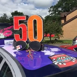 Decorative 50th birthday sign with sunglasses, a gold balloon, and firework confetti on the hood of a police car, with trees and buildings in the background.