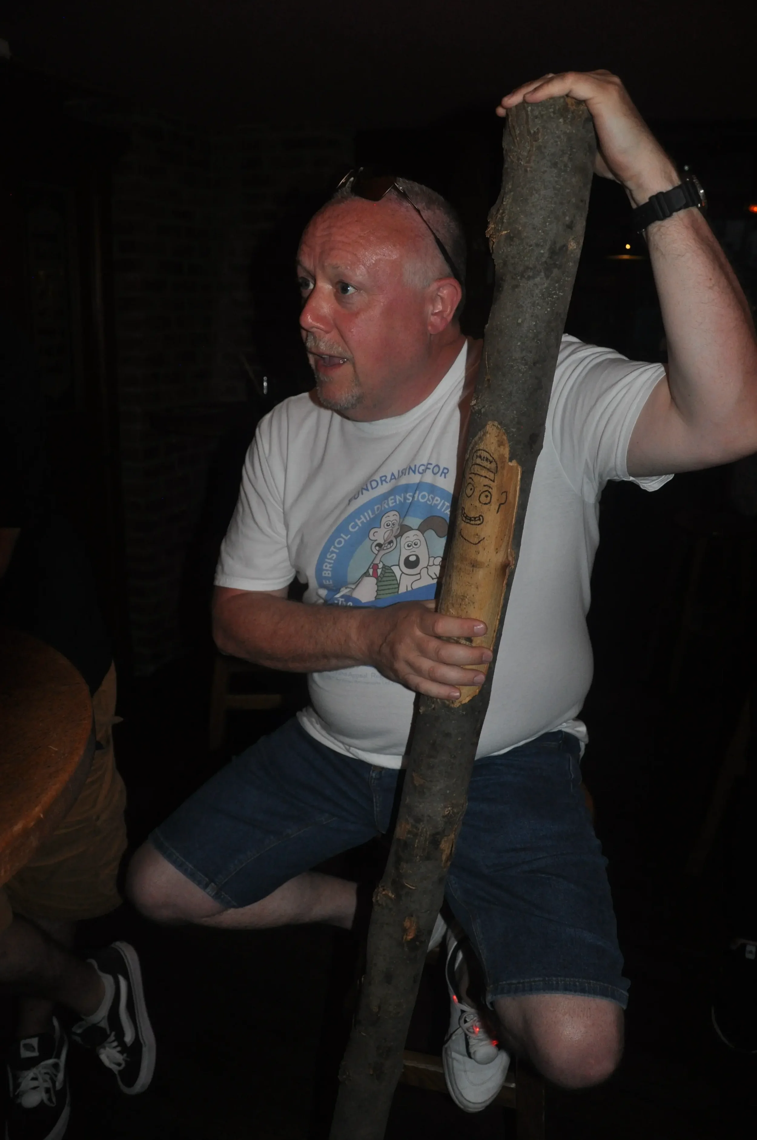 Man sitting on a stool holding a large tree branch with a face drawn on it, in a dimly lit room.