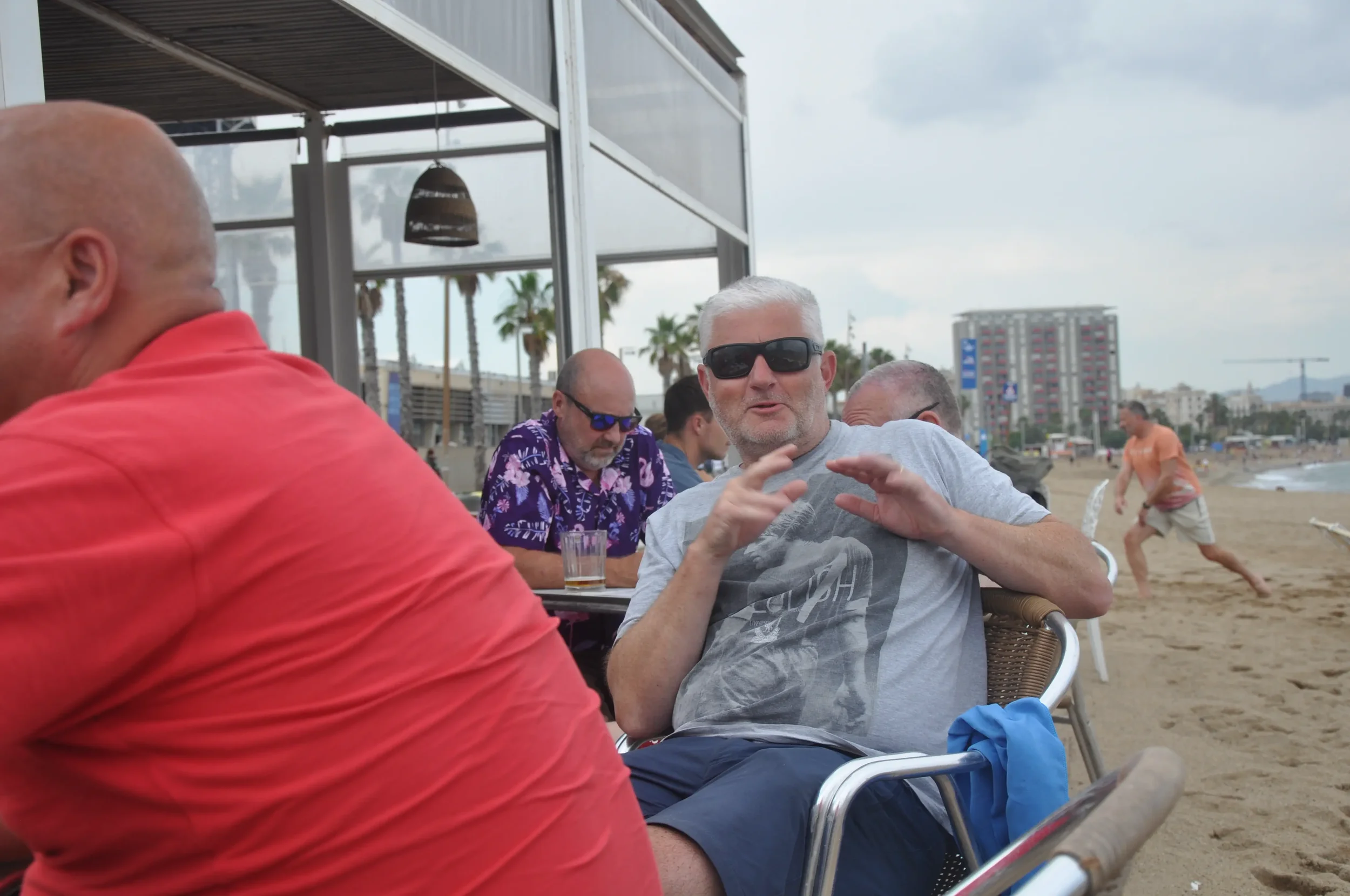 Group of men sitting outdoors at a beachside restaurant, with one man in sunglasses talking, and others around him, some with drinks. In the background, people are on the beach and walking along the shoreline, with buildings and palm trees.