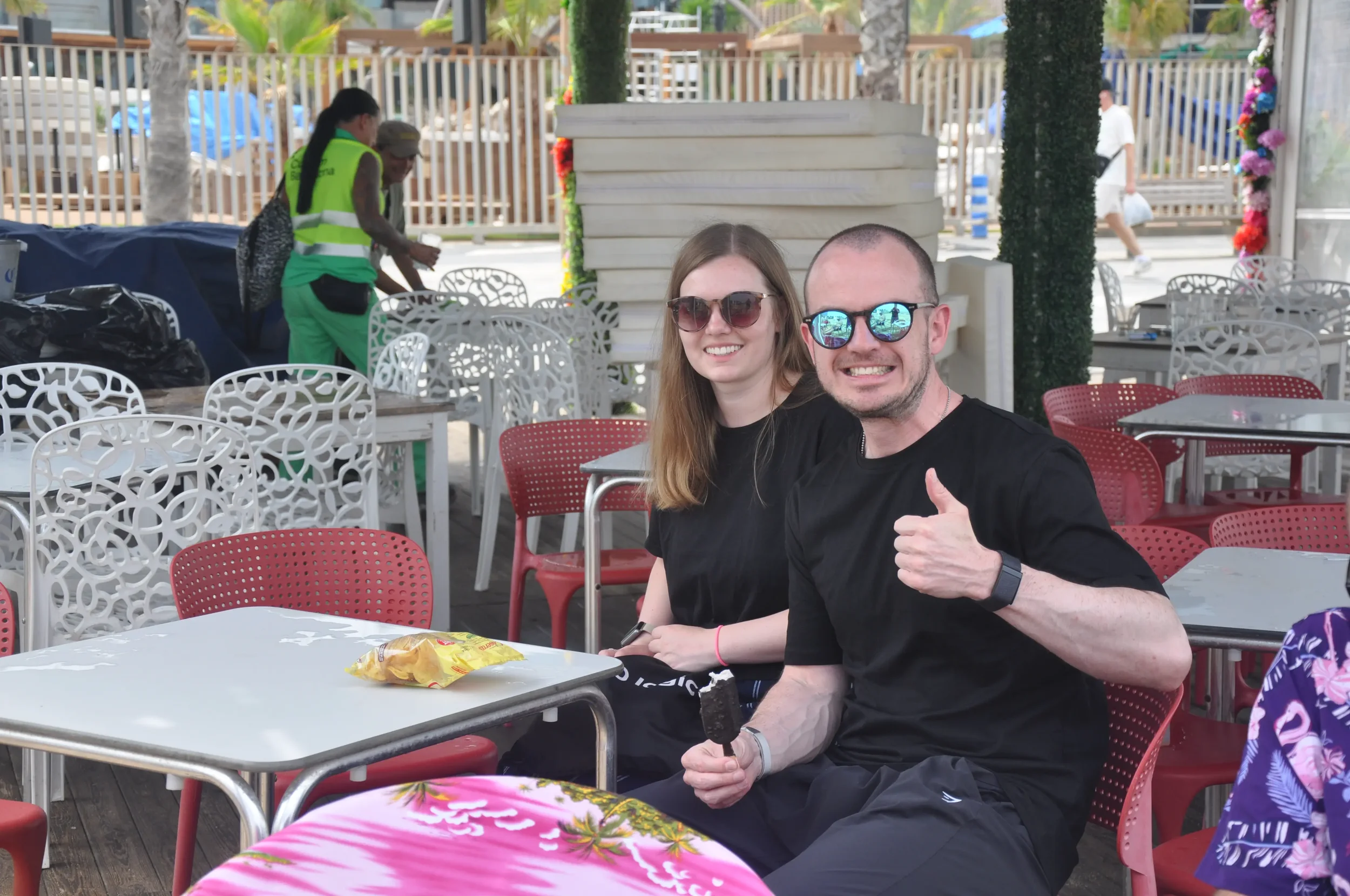 A smiling man with sunglasses giving a thumbs-up, holding a chocolate-covered ice cream bar, sitting next to a smiling woman also wearing sunglasses at an outdoor seating area with colorful chairs. Behind them, there are two women standing and talkin