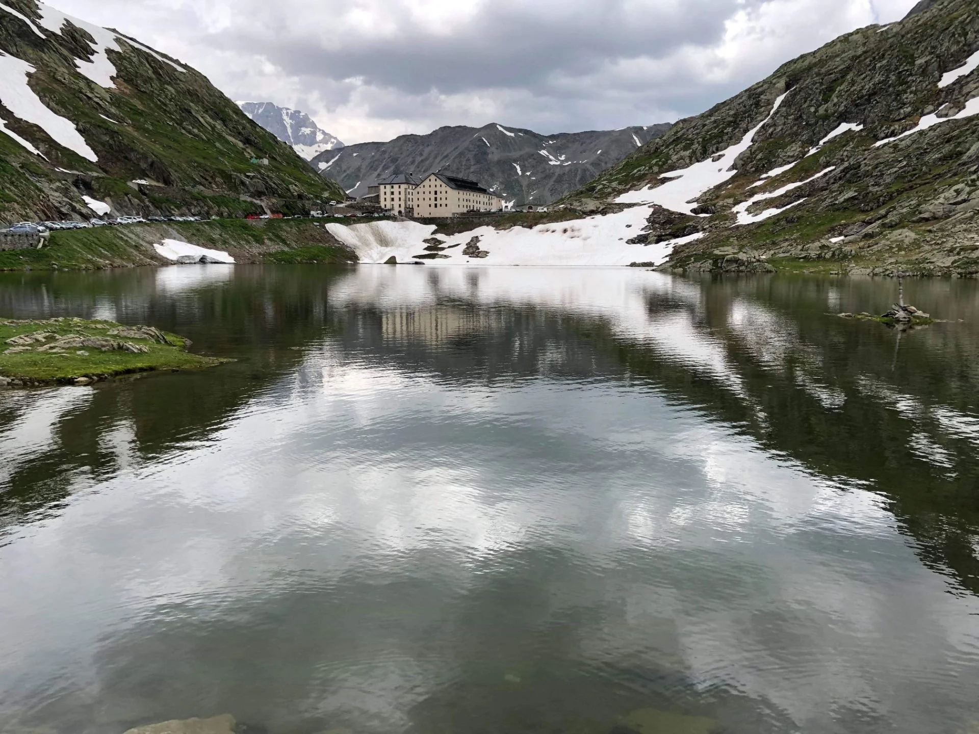 Mountain lake surrounded by snow-capped hills and buildings in the background, with cloudy sky reflected in the water.