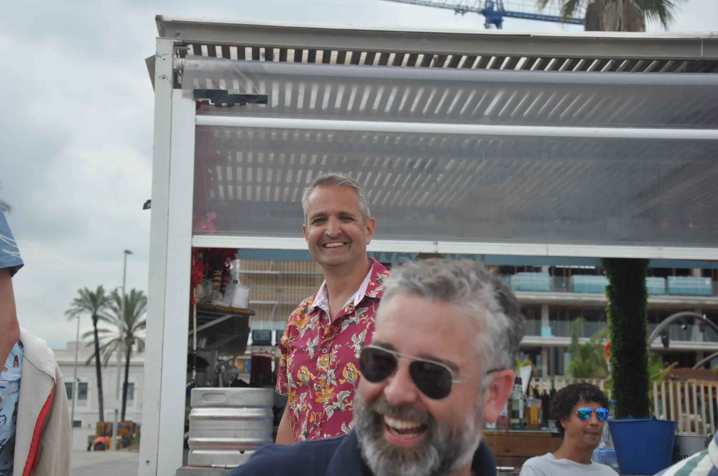 Group of four people enjoying outdoors with palm trees and modern buildings in the background, one man in a colorful hawaiian shirt smiling, another man with gray hair and beard wearing sunglasses laughing, a woman with curly hair wearing blue sungla