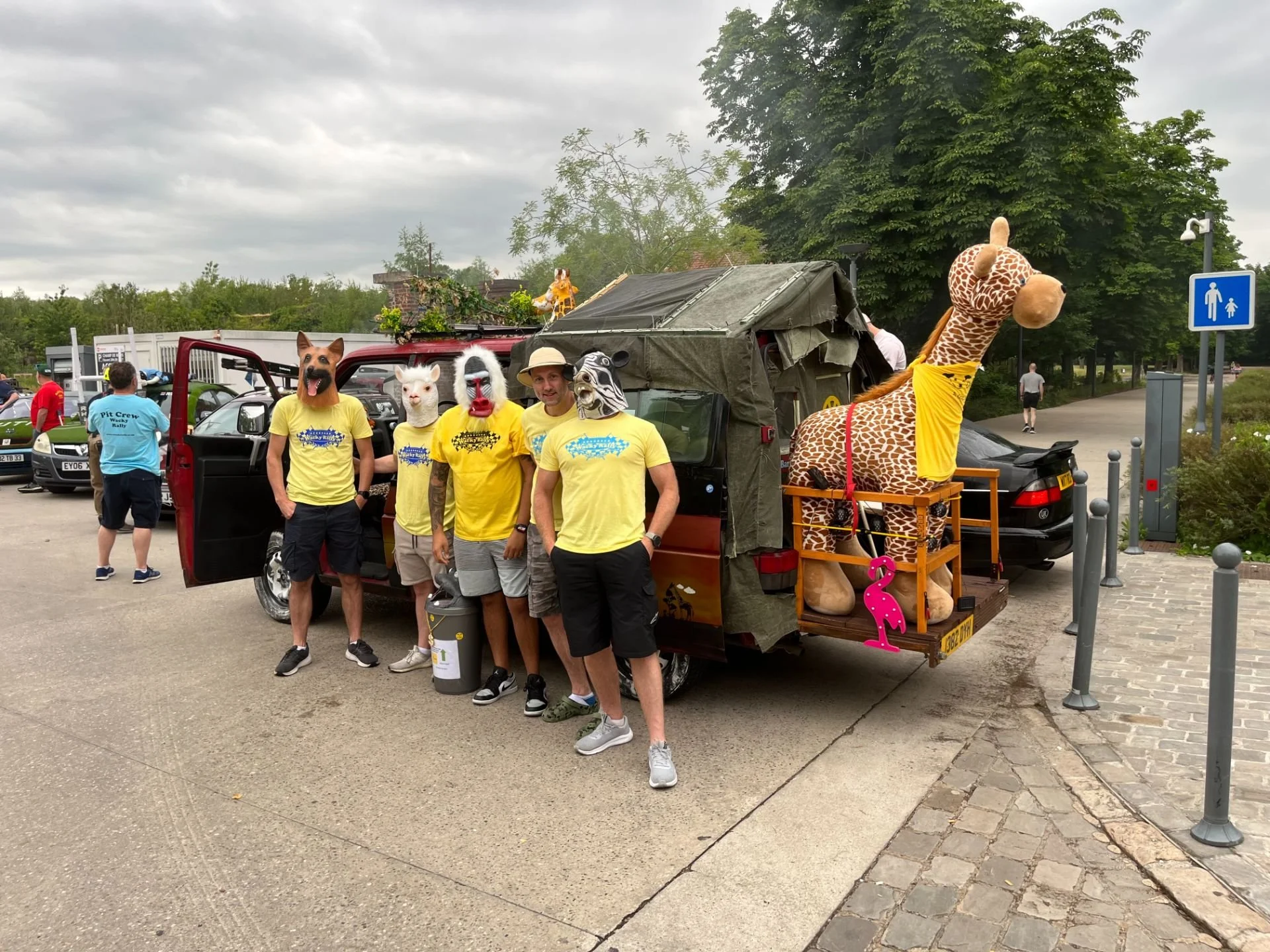 Four people wearing yellow t-shirts with animal masks standing next to a decorated vehicle with a large giraffe figure on the back, in an outdoor parking area.