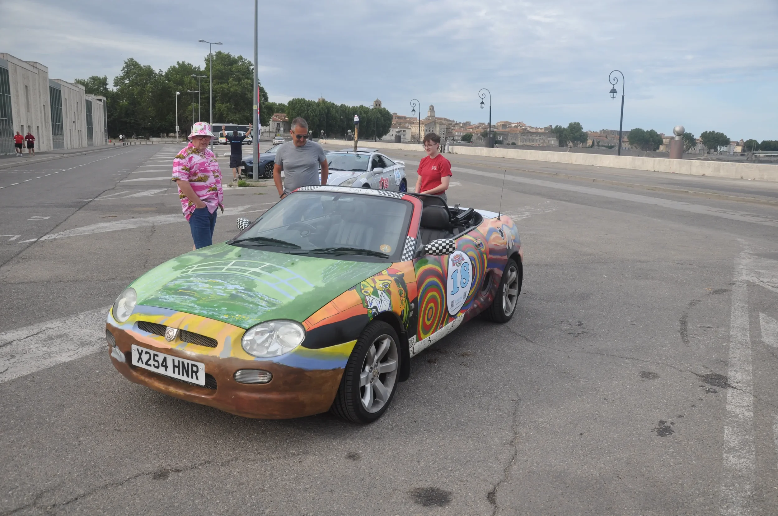 A colorful race car with artwork and the number 18 on its side, parked in an empty parking lot with a few people standing nearby and a scenic cityscape in the background.