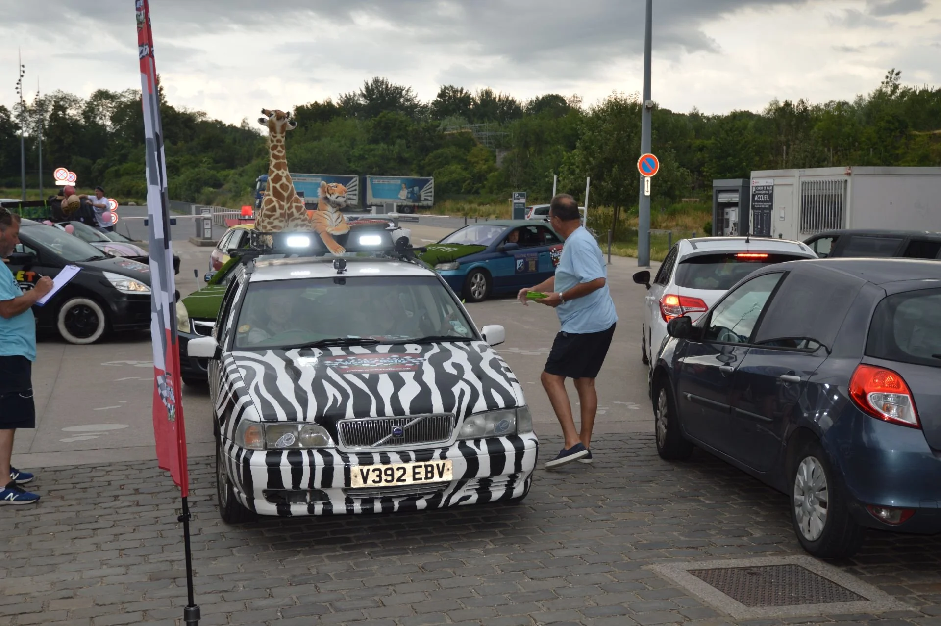 Parking lot with cars, a man is checking in with a person, a car with zebra stripes and a giraffe and tiger plush toys on the roof.