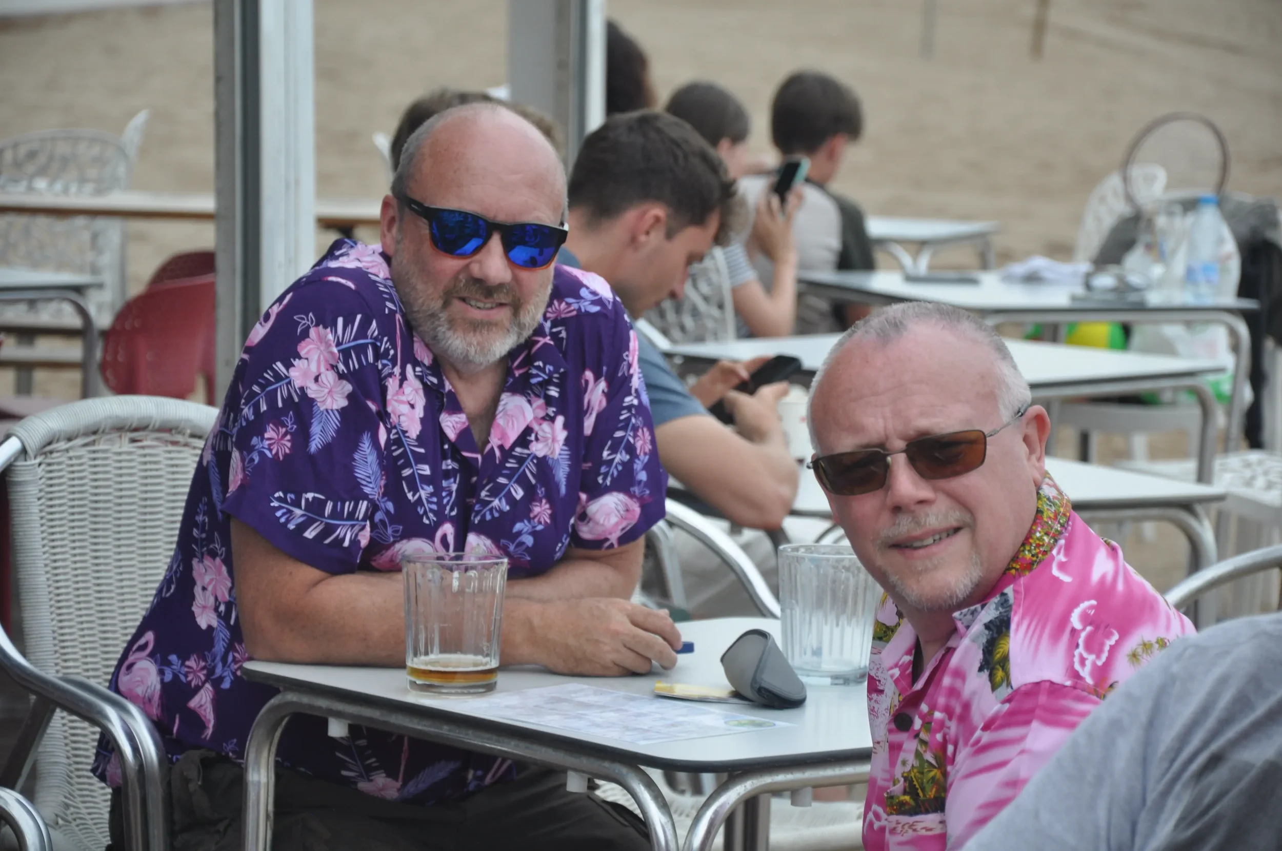 Two middle-aged men wearing colorful Hawaiian shirts and sunglasses sitting at an outdoor table with drinks, smiling at the camera. Other people are seated at tables in the background.