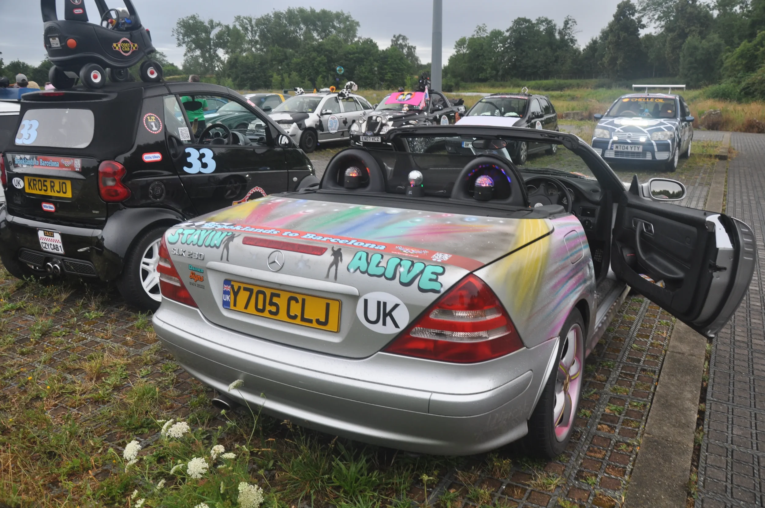 Various decorated cars parked outdoors on a gravel and paved area, with some cars featuring stickers, artwork, and accessories.