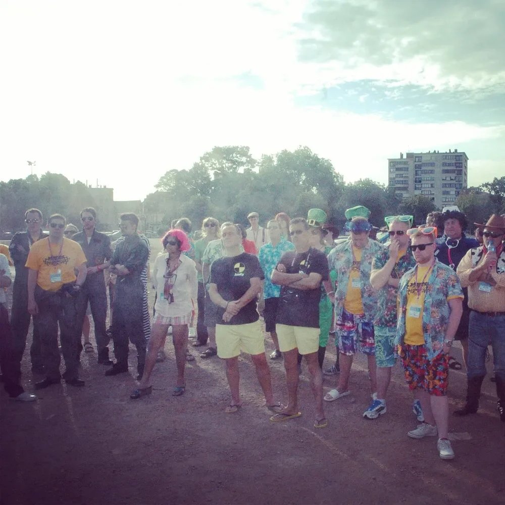 Group of people dressed in colorful, tropical and costume attire, gathered outdoors with a cityscape and trees in the background.