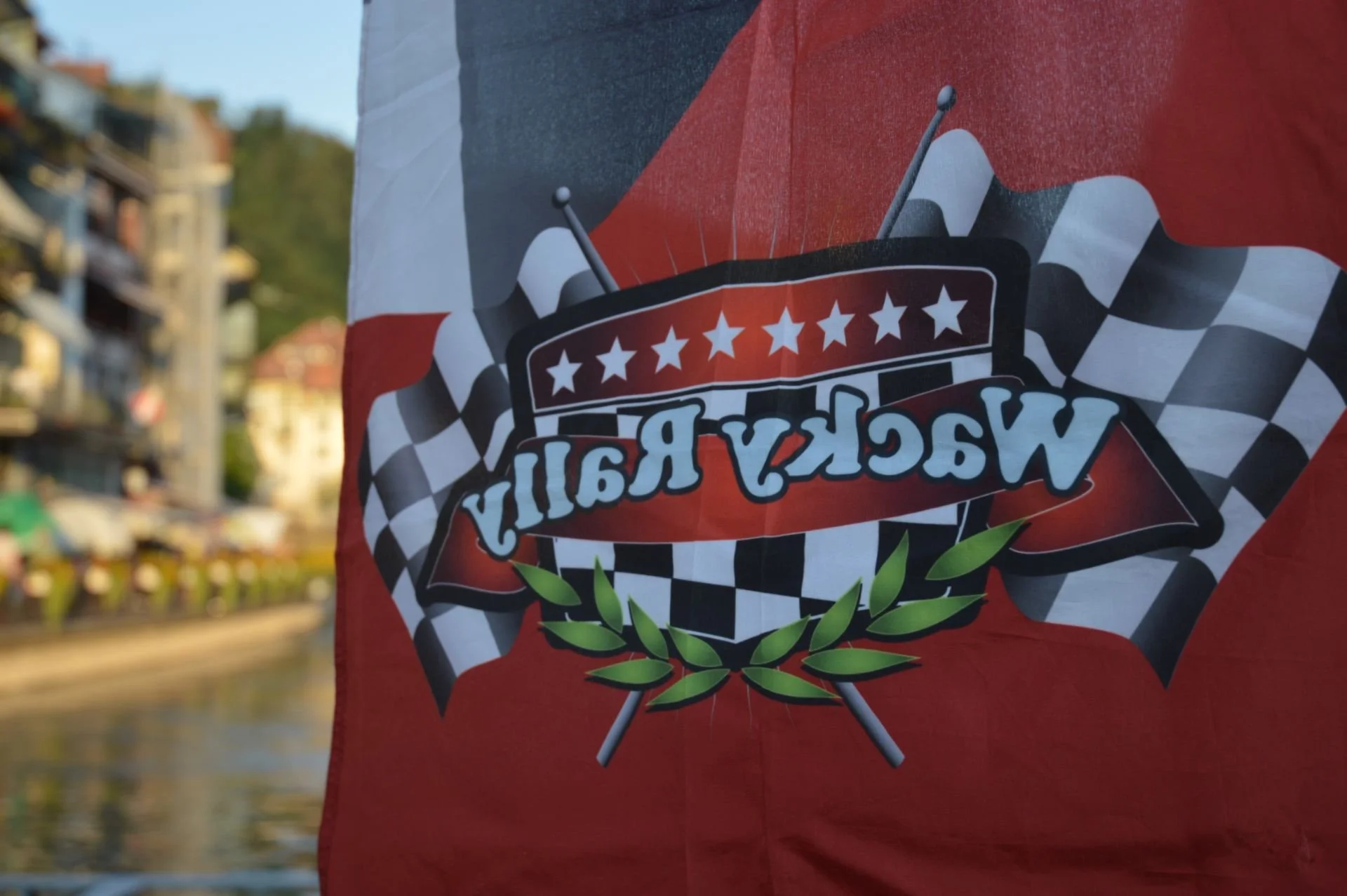 A flag with a racing theme, featuring checkered black and white flags, red background, and a emblem with stars, laurel leaves, and the text "Race Now" upside down.