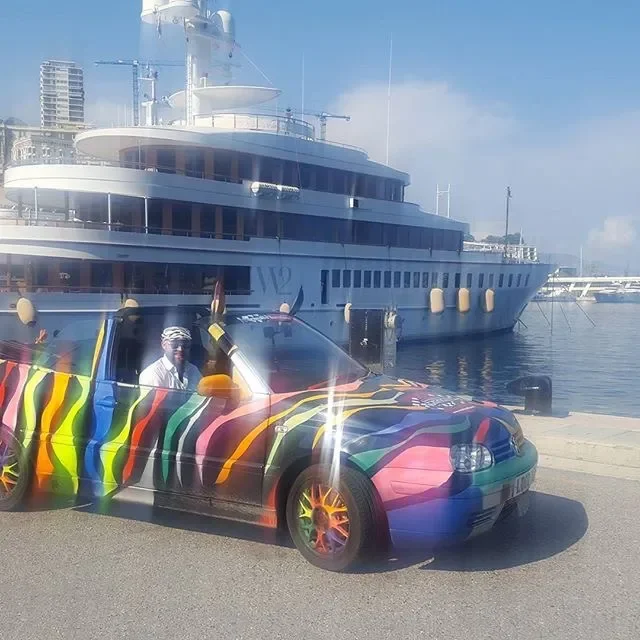 Colorful sports car with rainbow paint, parked near a large yacht at a marina.
