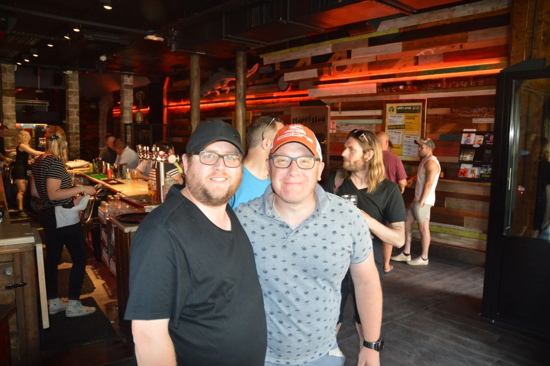 Two men smiling at the camera in a busy bar with people socializing in the background.