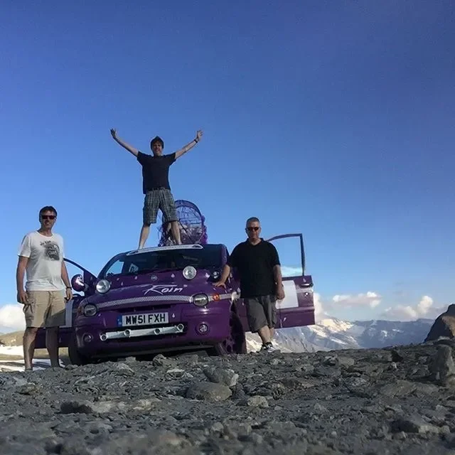 Three men standing on rocky terrain with one person standing on top of a purple car with arms raised, mountains in the background, clear blue sky.