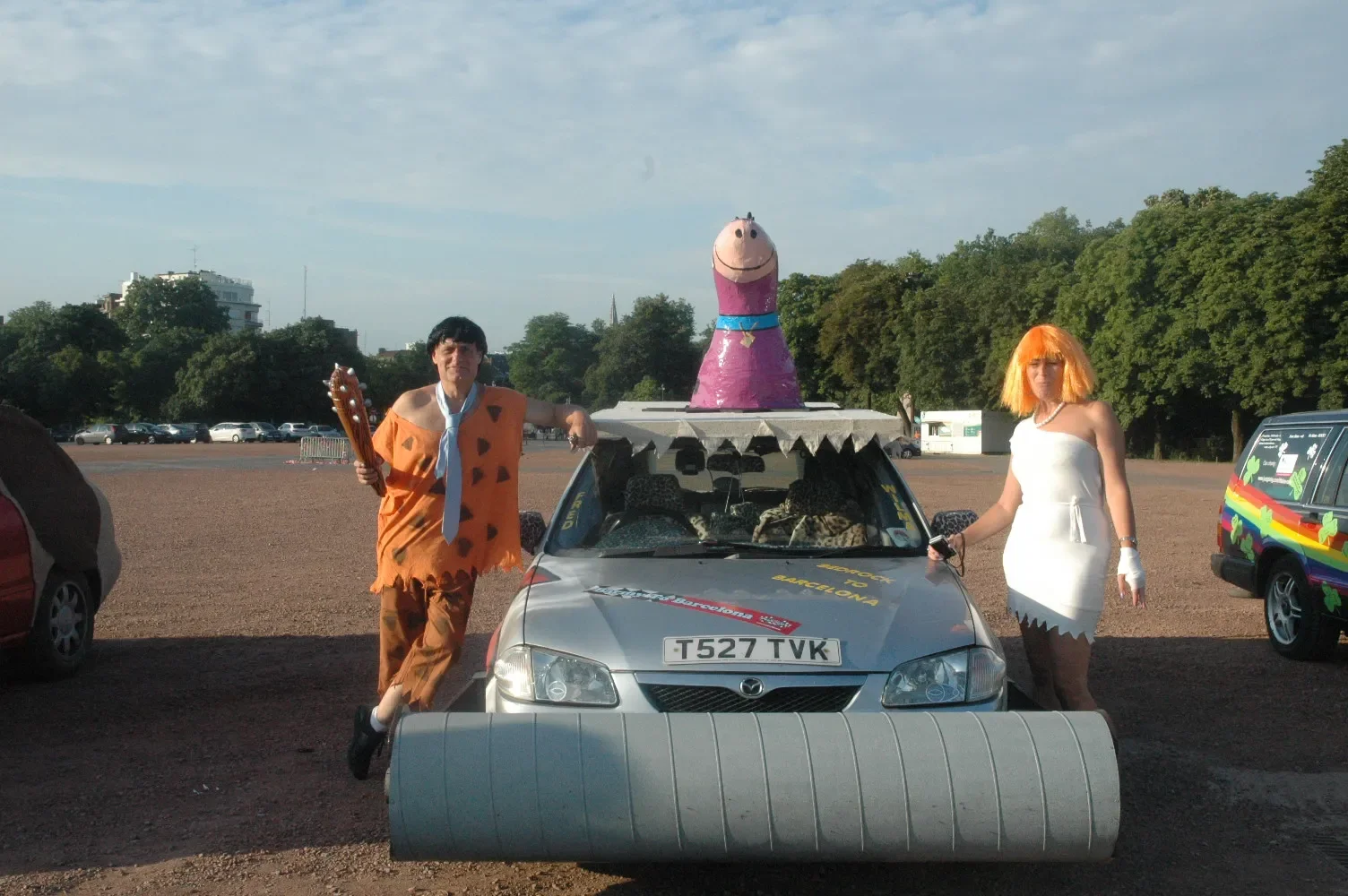 Two people dressed as cartoon characters stand next to a modified car in a parking lot, with a large pink cartoon dog head on top of the car and a foam roller in front of the car.