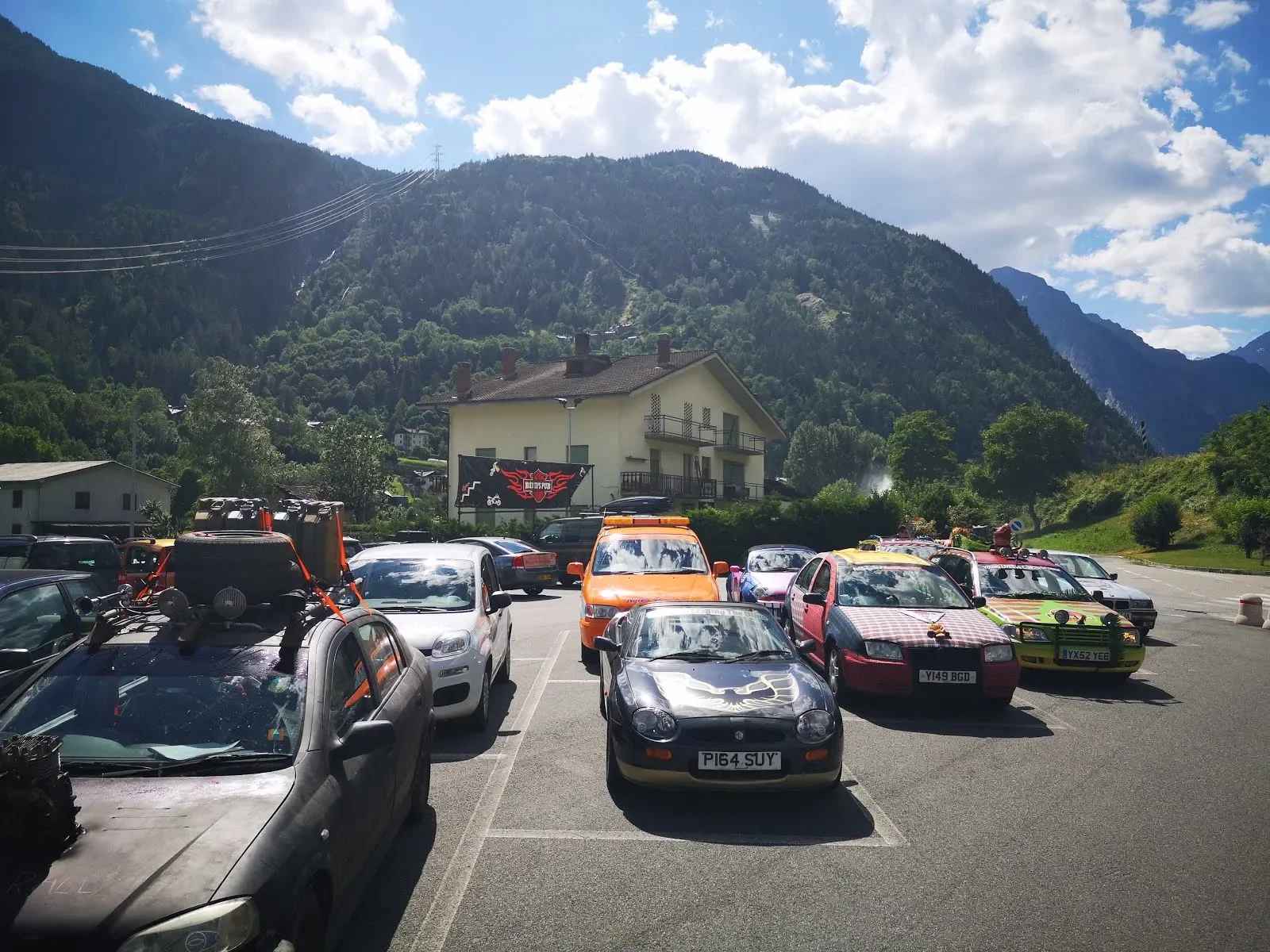 A parking lot filled with various cars in front of a house, mountains, and a partly cloudy sky.