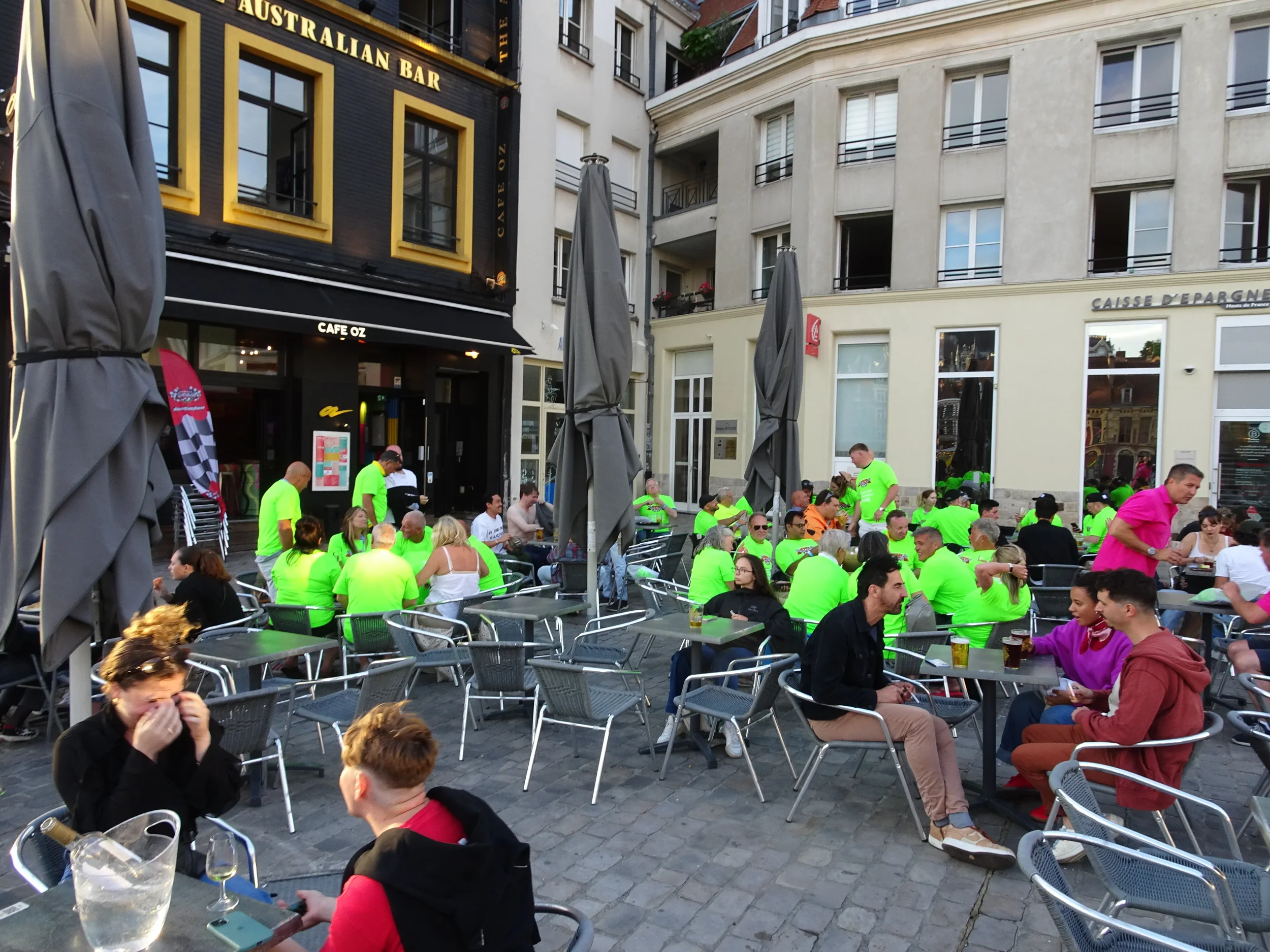 People sitting at outdoor tables in a busy city plaza, some wearing neon green shirts, others in casual clothing. The scene shows a lively atmosphere outside a bar and restaurant with umbrellas and building facades in the background.