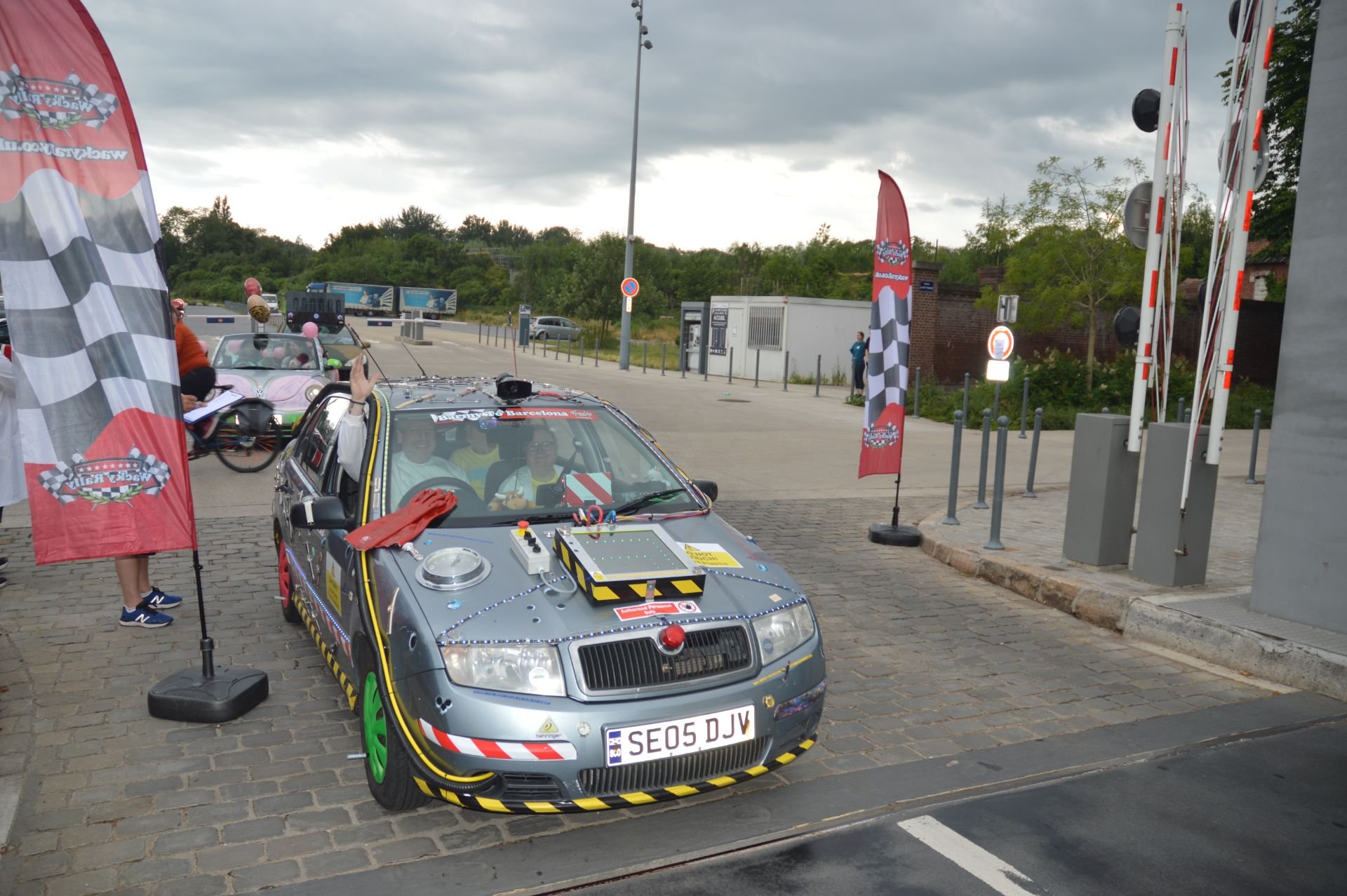 A street scene with a decorated car resembling a crash test vehicle, participating in a rally event, with flags and people in the background under cloudy skies.