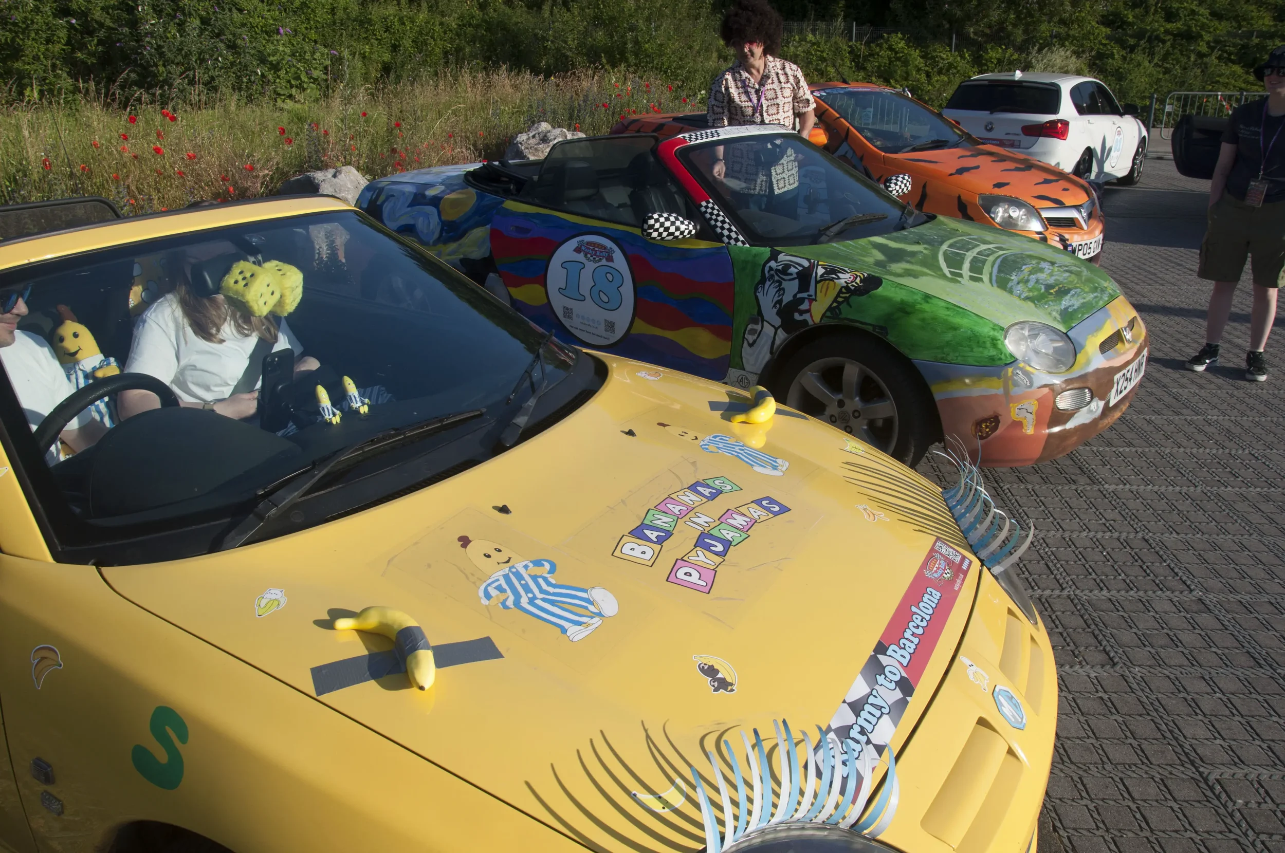 Colorful convertible cars decorated with cartoon characters and travel-themed stickers are parked next to each other. There are people and a woman in sunglasses standing nearby, with a grassy area and flowers in the background.
