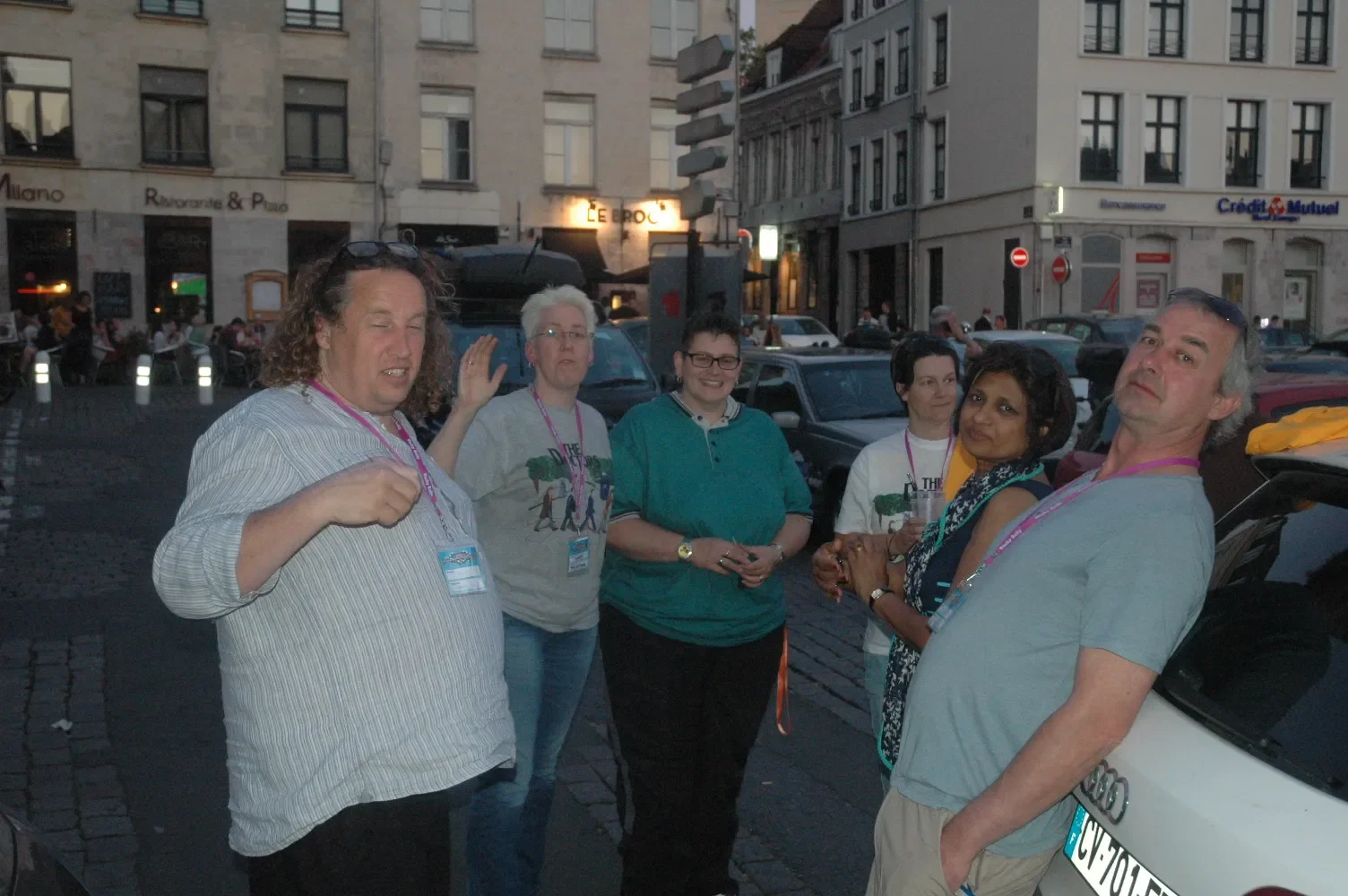 A group of six diverse people standing on a city street in the evening, with buildings and cars in the background, some wearing badges and casual clothing, engaged in conversation.