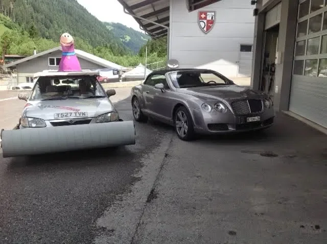 A silver car with a large foam roller attached to its front bumper, parked next to a gray luxury convertible car, outside a building with a Swiss shield emblem on the wall. In the background, there is a colorful inflatable figure and green hills.