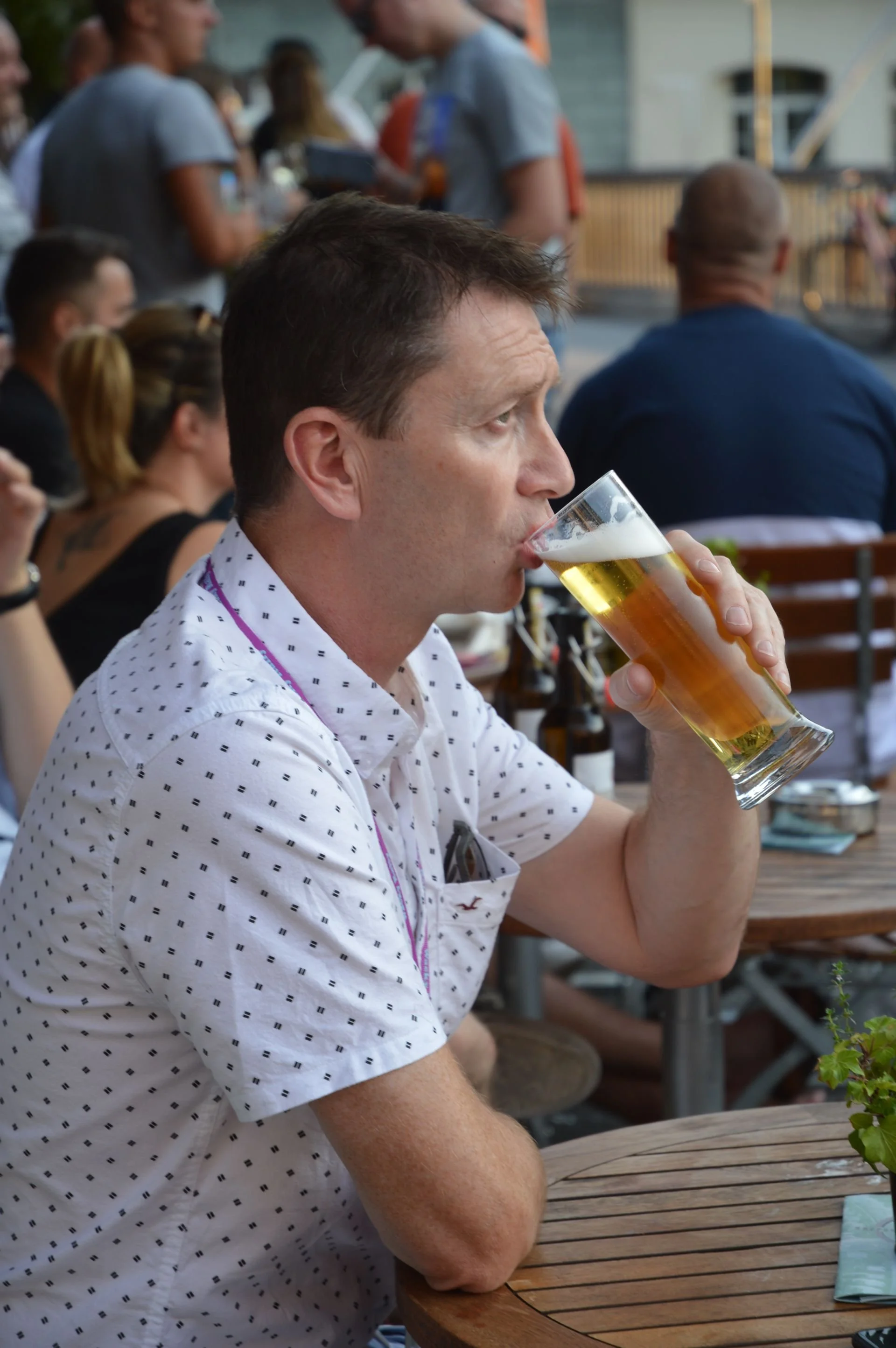 A man sitting at an outdoor table drinking a glass of beer, surrounded by people at a social gathering.